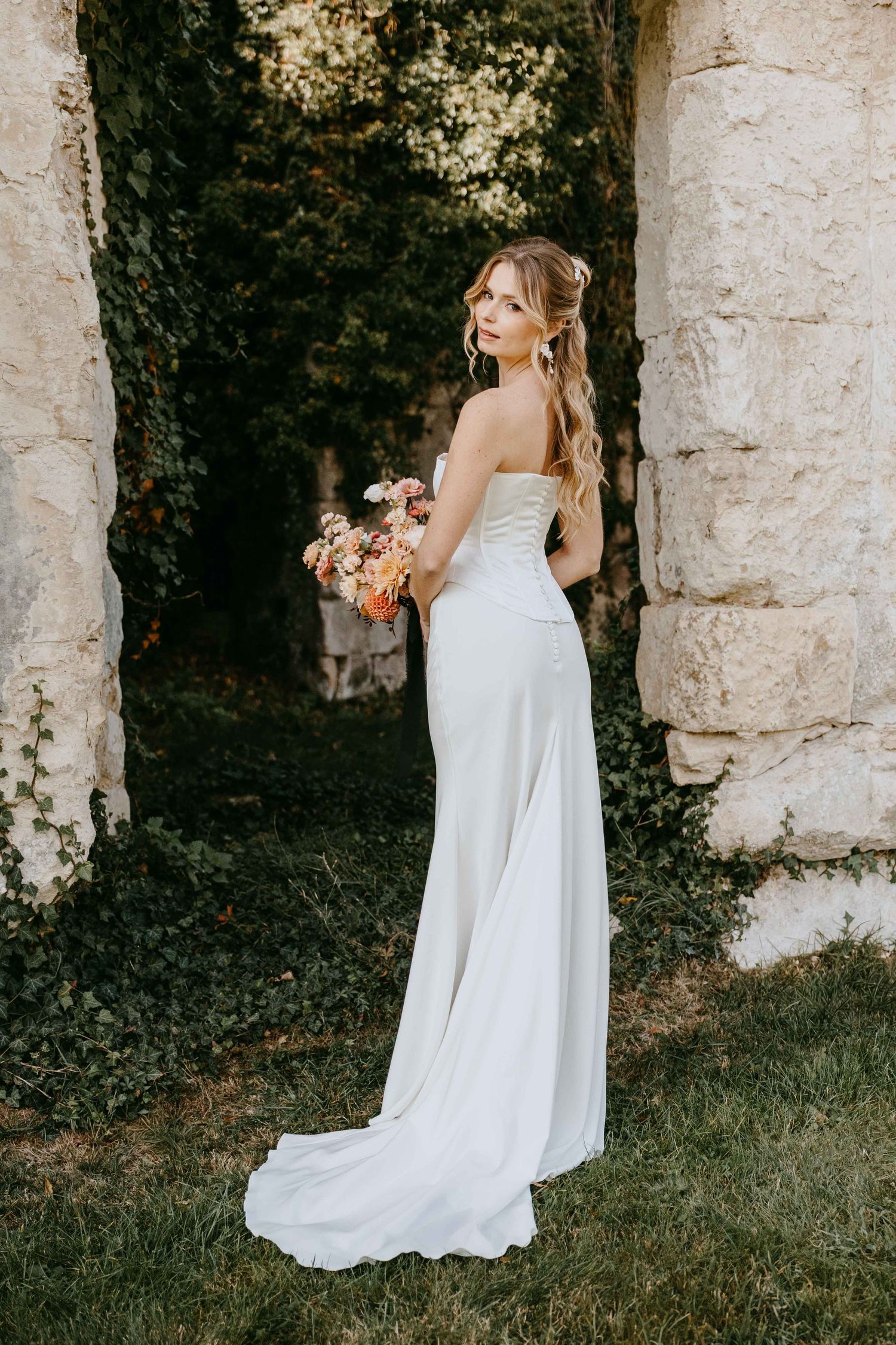 Bride in strapless white gown holds colorful bouquet, posing gracefully between stone columns with lush ivy backdrop.