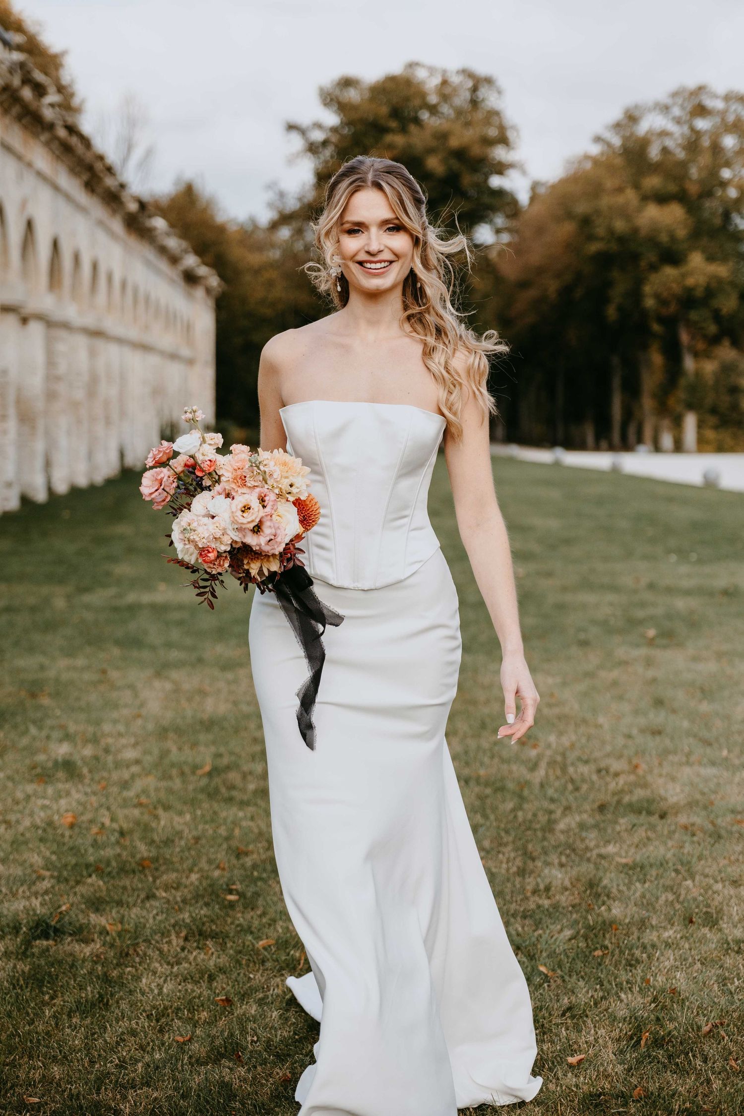 Smiling bride in strapless white gown holding colorful bouquet with black ribbon, posing outdoors near stone arches.
