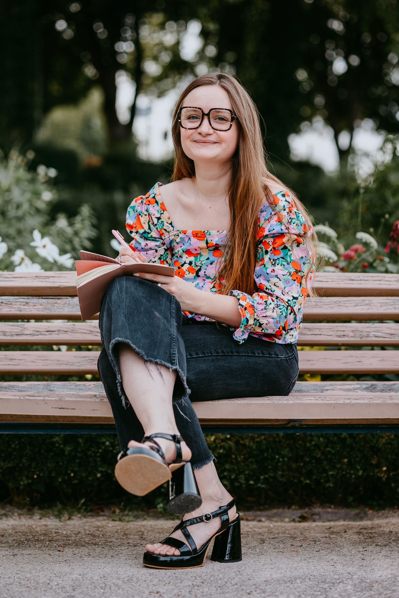 Young woman with glasses reading a book on a park bench, wearing a floral blouse and jeans.