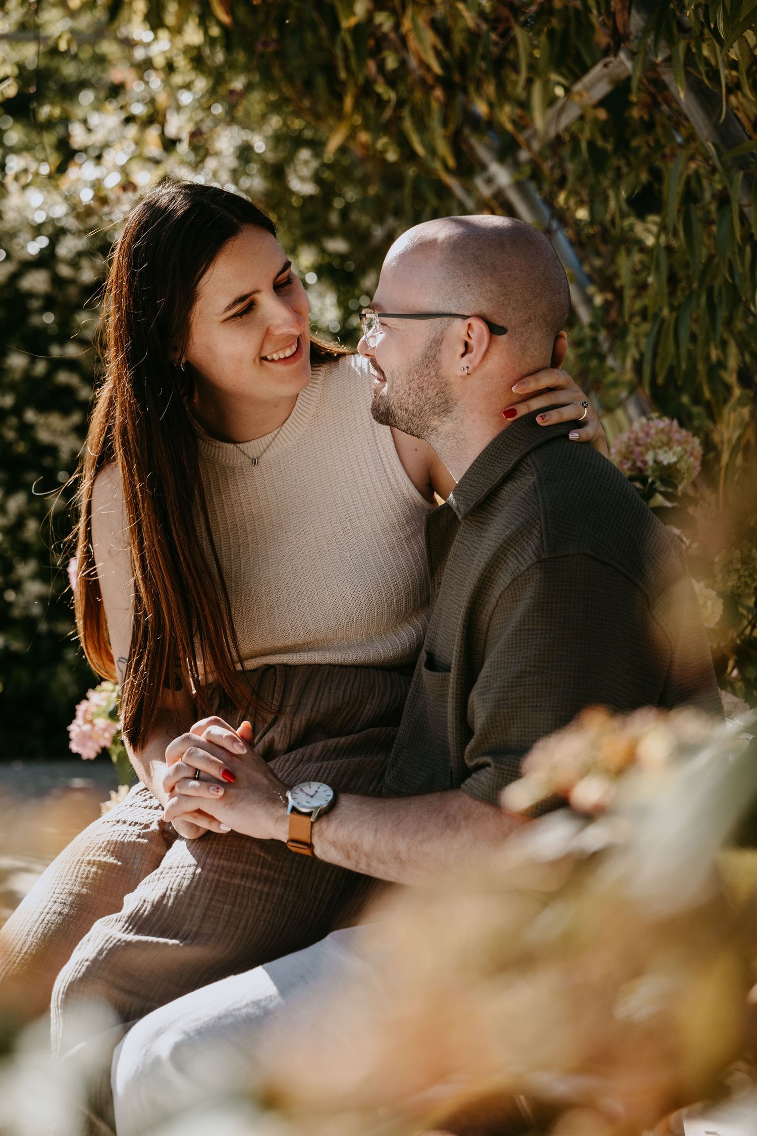 A couple sitting outdoors, smiling and holding hands, surrounded by lush greenery in warm natural light.