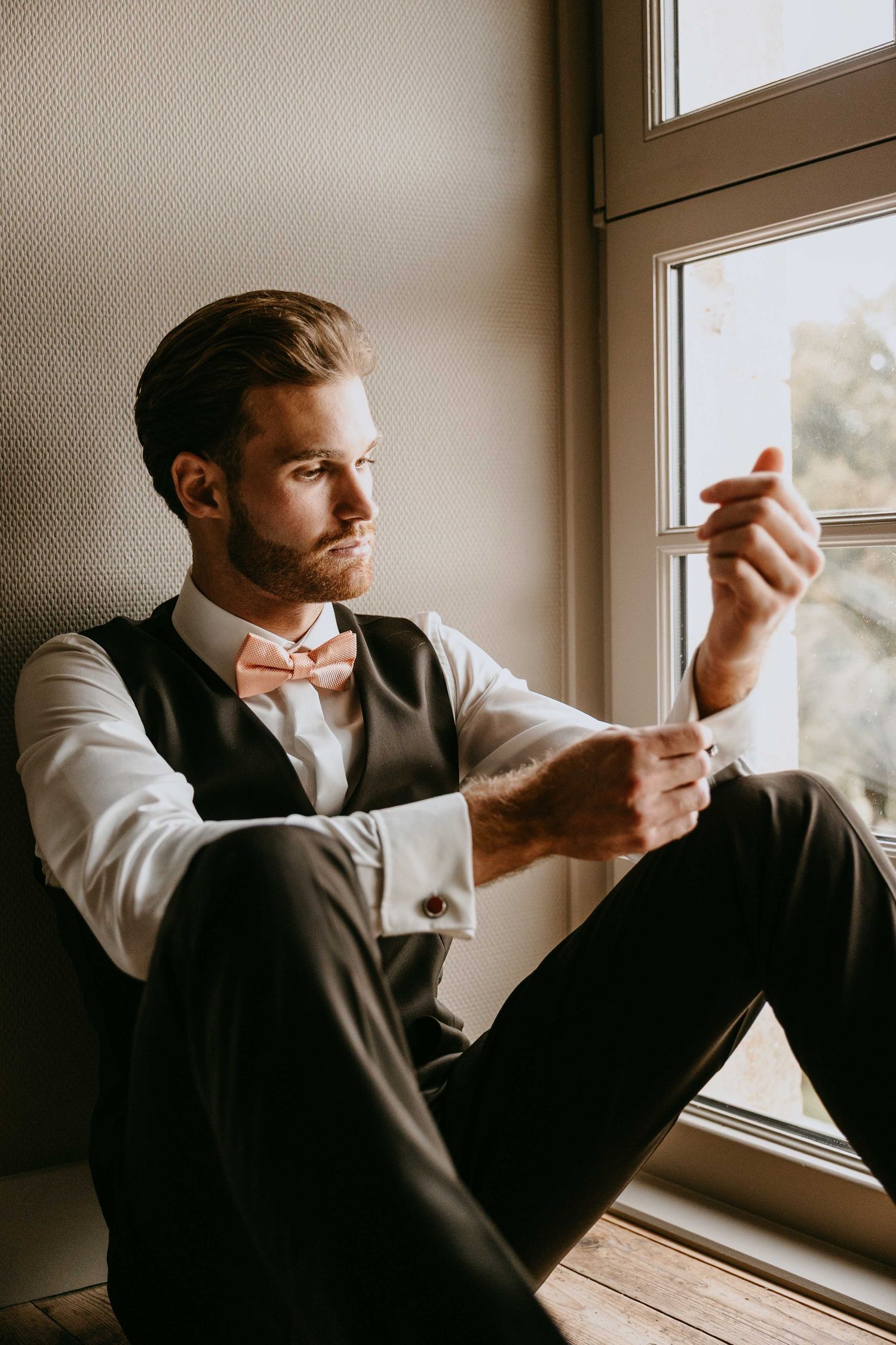 Dapper groom in vest and pink bow tie sitting by window examining cufflinks before wedding ceremony.