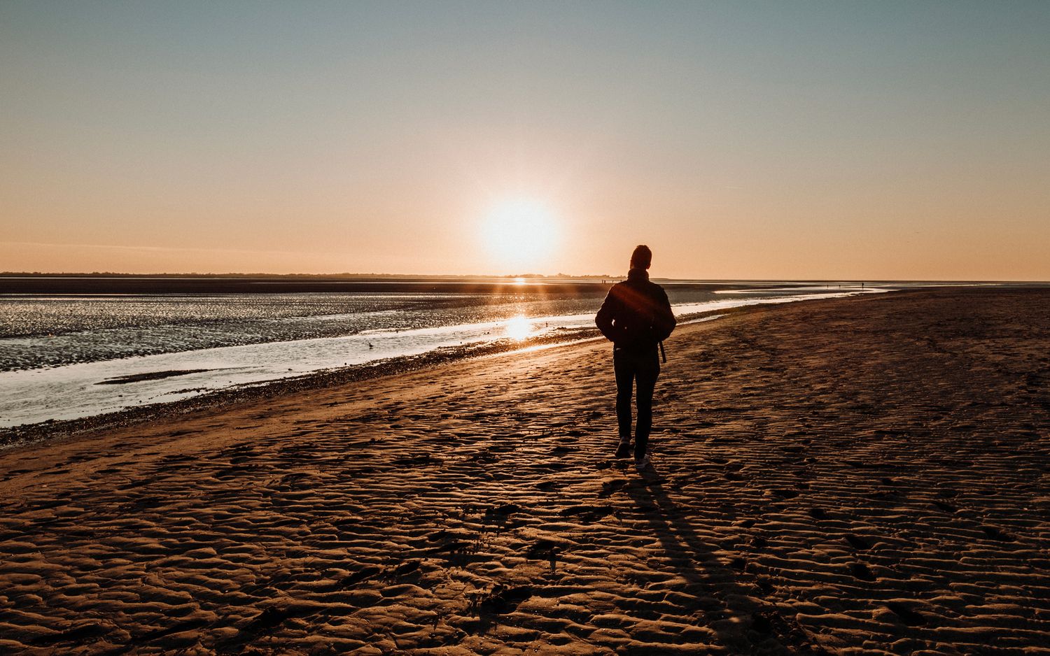 Silhouette of a person walking alone on a vast sandy beach during a golden sunset.