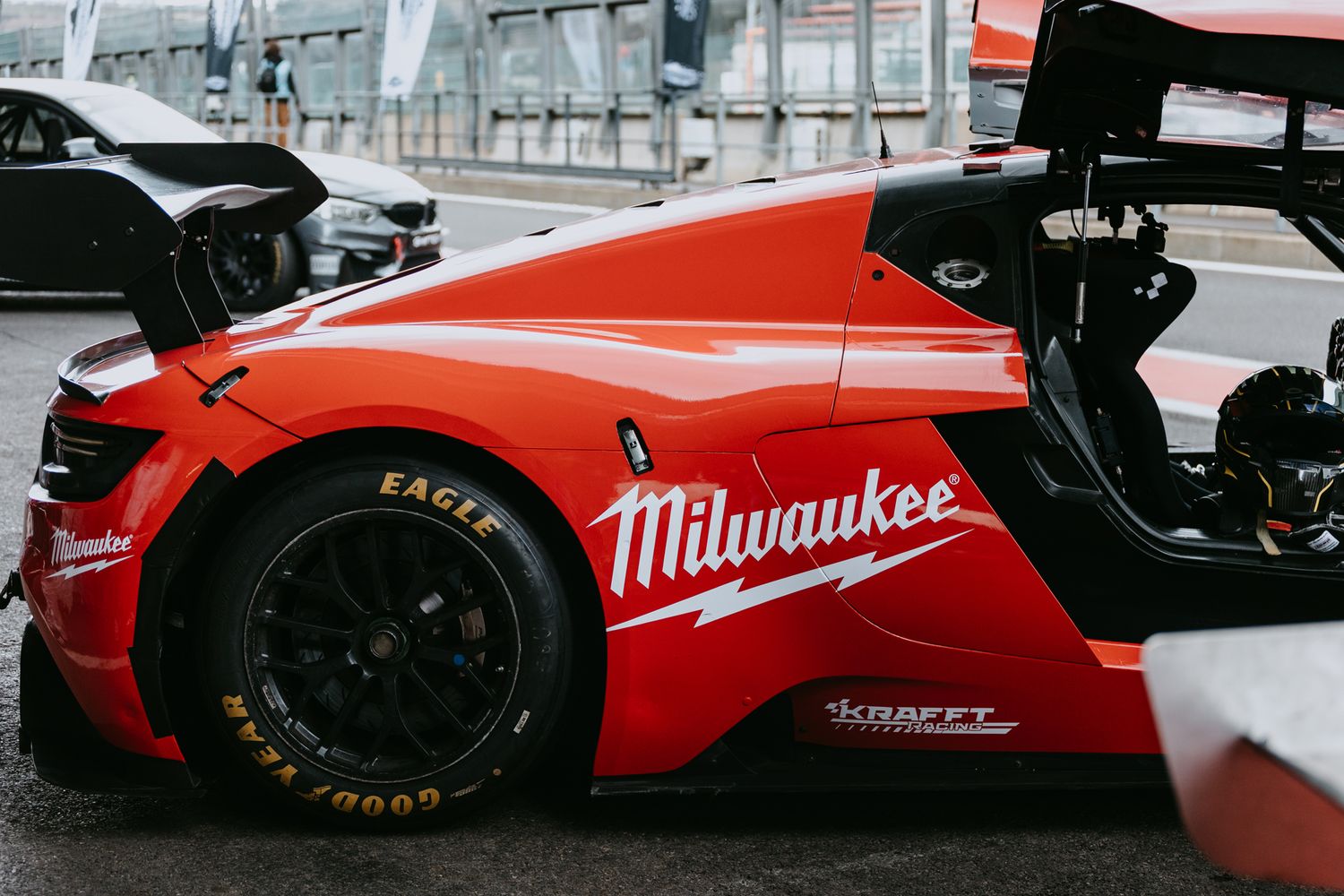 Red Milwaukee-branded racing car with open door parked on a race track pit lane.