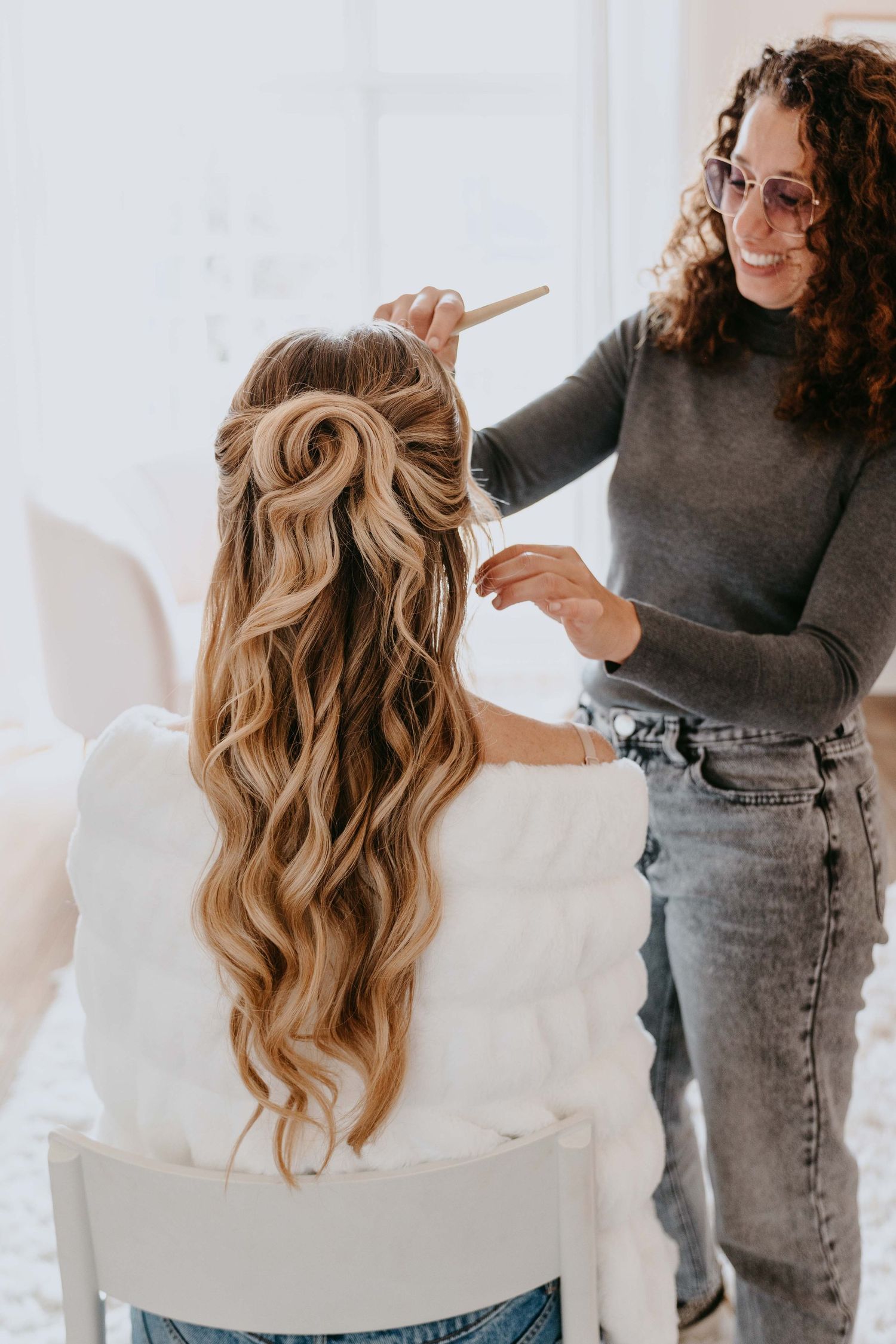 Hair stylist with curly hair styling long blonde wavy hair in a half-up style on a seated client.
