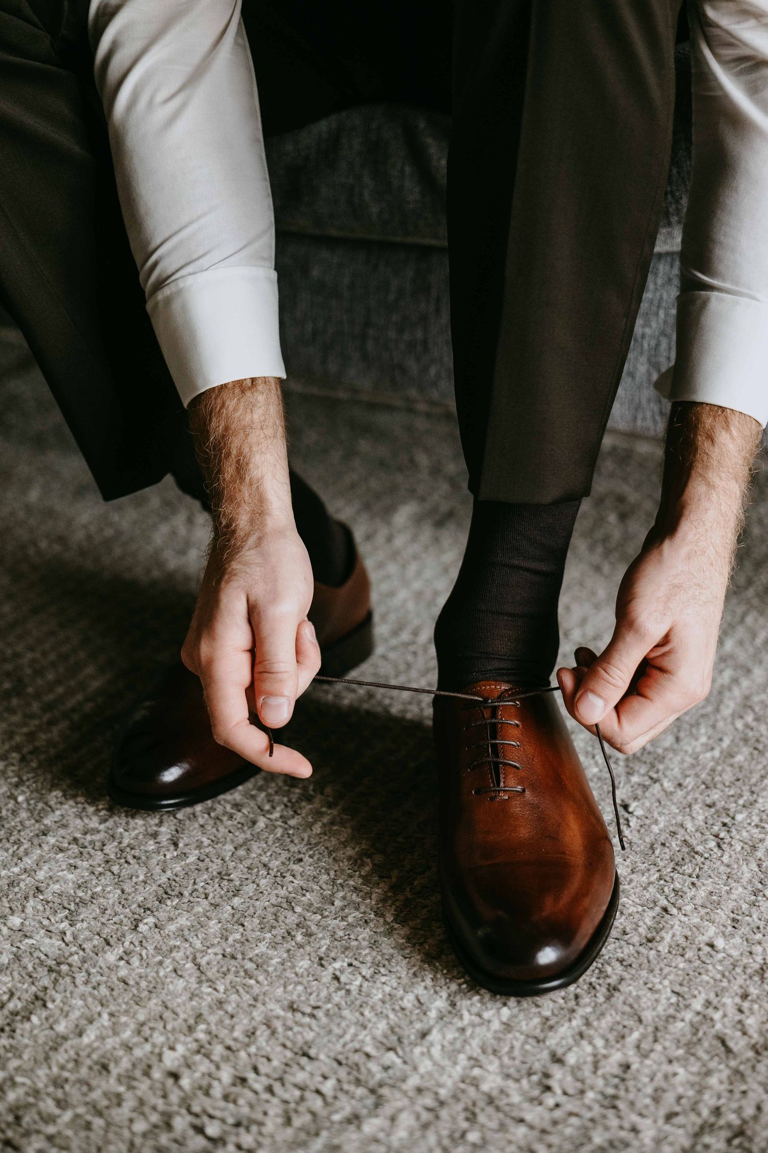 Groom tying laces on polished brown leather Oxford dress shoes on wedding day.