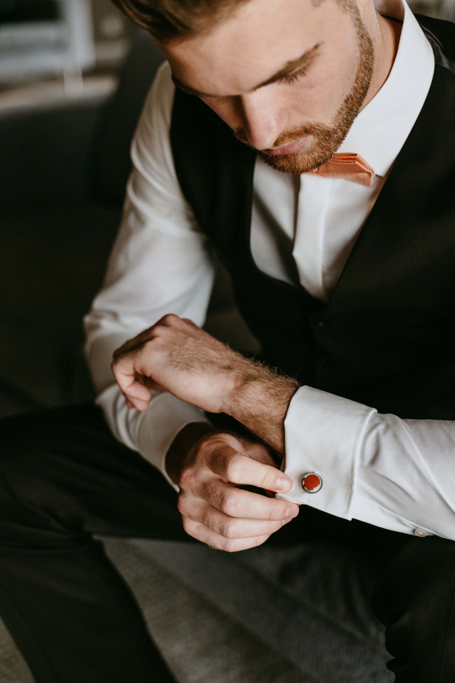 A well-dressed groom in a black vest and white shirt fastens a red cufflink while preparing for his wedding day.