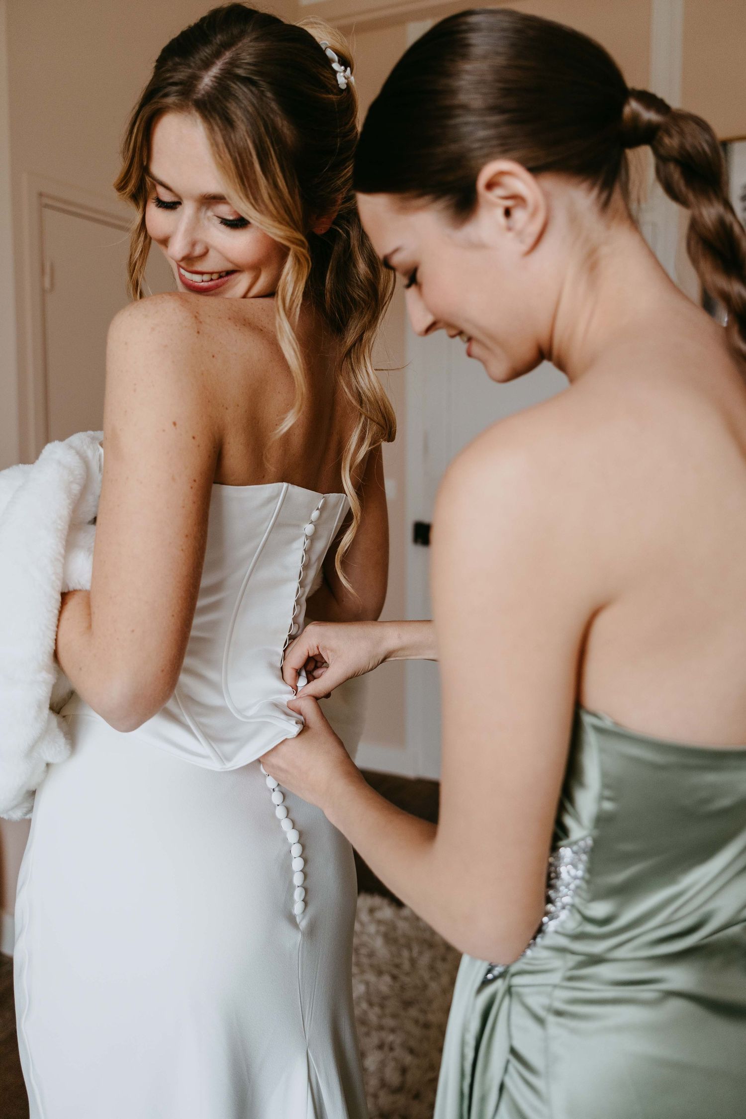 Bridesmaid in sage green dress fastening buttons on bride's white strapless wedding gown during bridal preparations.