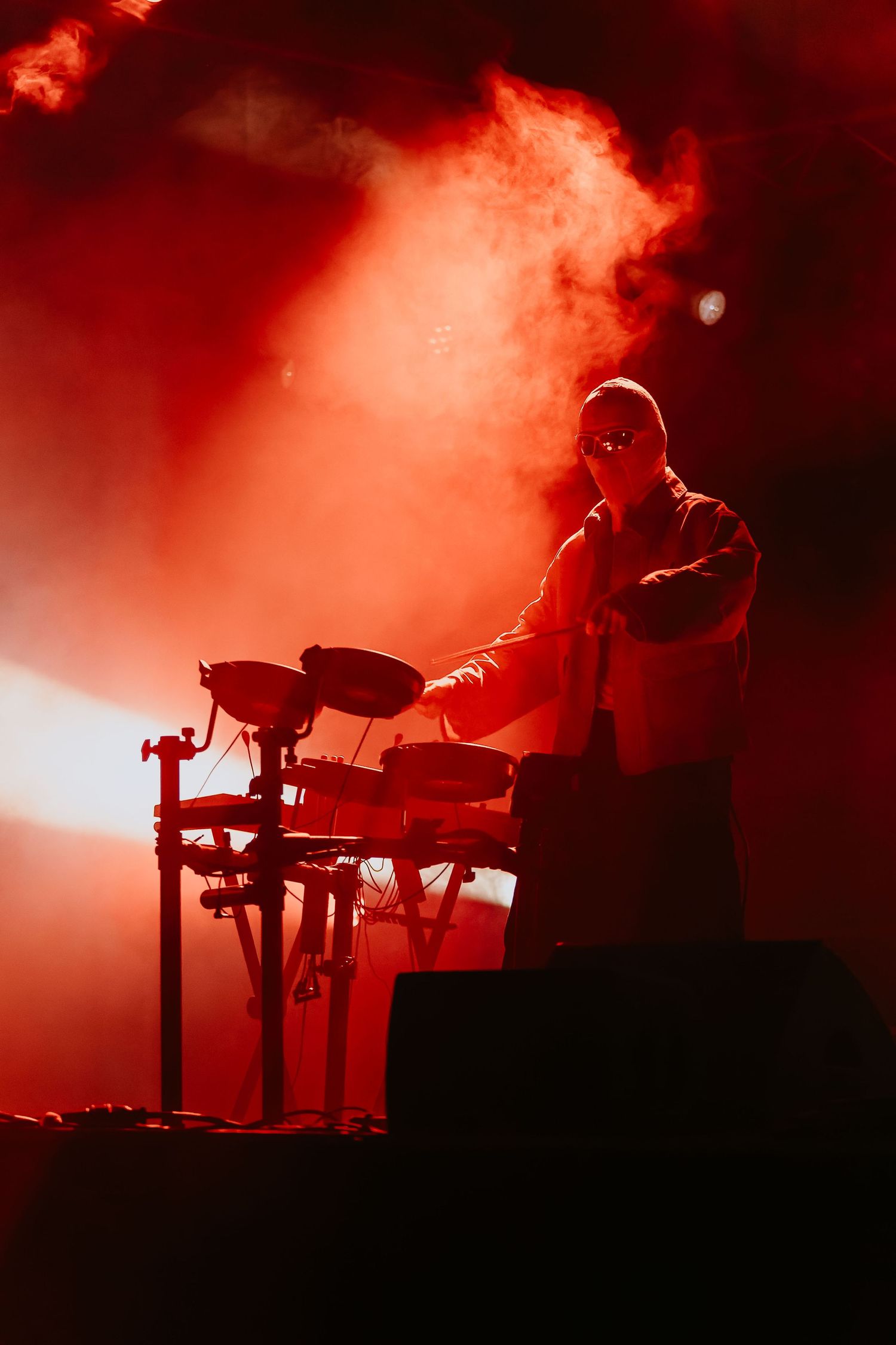 Silhouetted drummer performing at an electronic drum kit on a smoky red-lit concert stage.