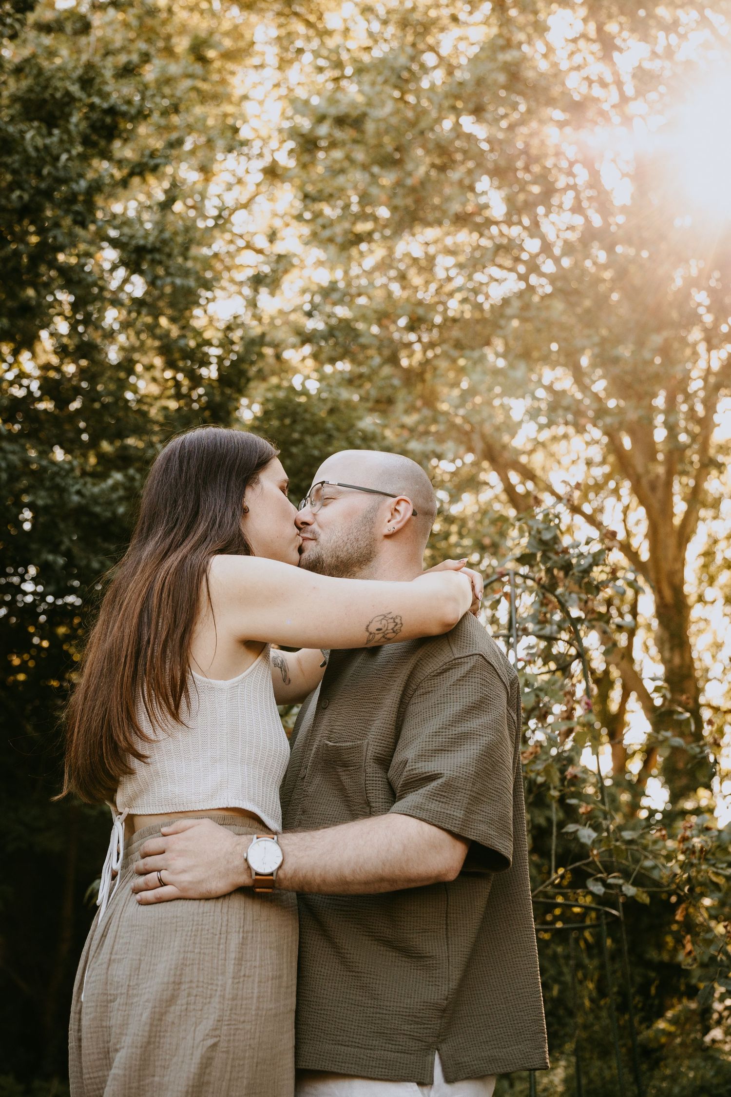 A couple sharing a tender kiss outdoors, embracing in golden sunlight with lush green trees in the background.