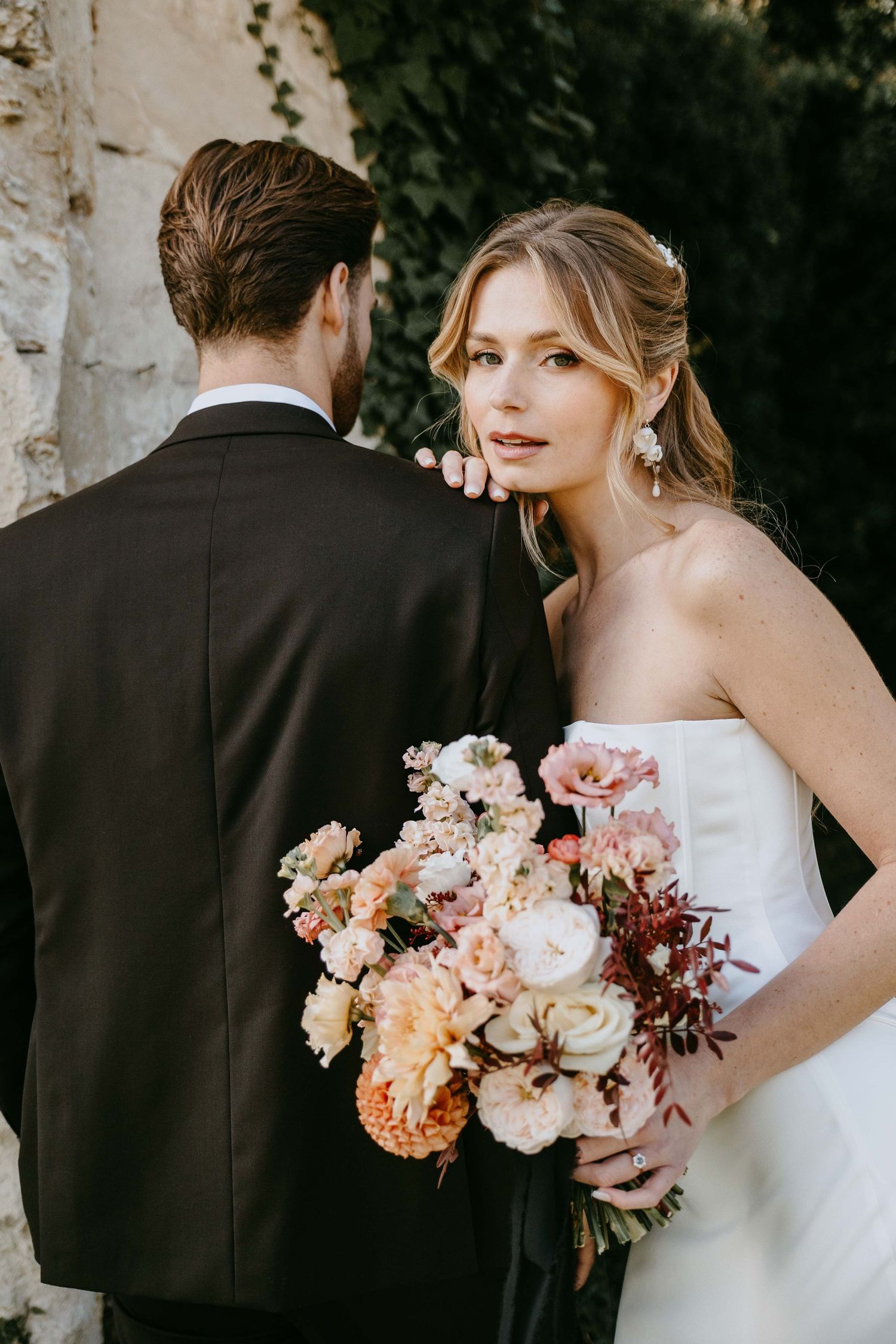 Elegant bride holding a pastel bouquet stands beside groom in black suit near stone wall.