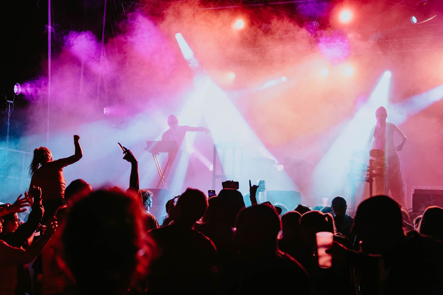 Energetic concert crowd cheering under colorful pink and purple stage lights with smoke effects.