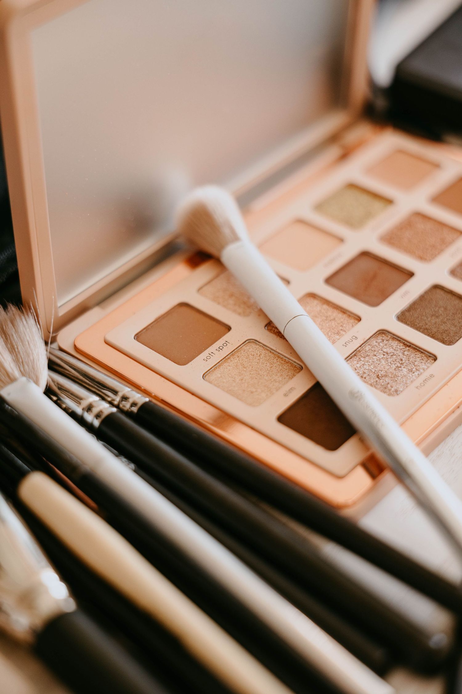 Neutral eyeshadow palette with warm tones, white makeup brush resting on top, surrounded by professional makeup brushes.