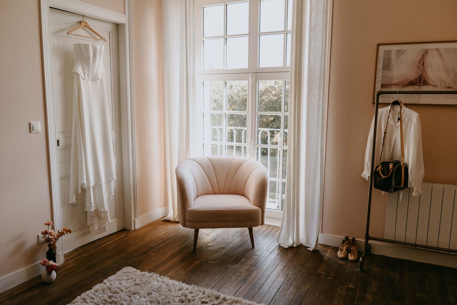 Elegant bridal suite with white wedding dress hanging, beige armchair, and natural light from French windows.