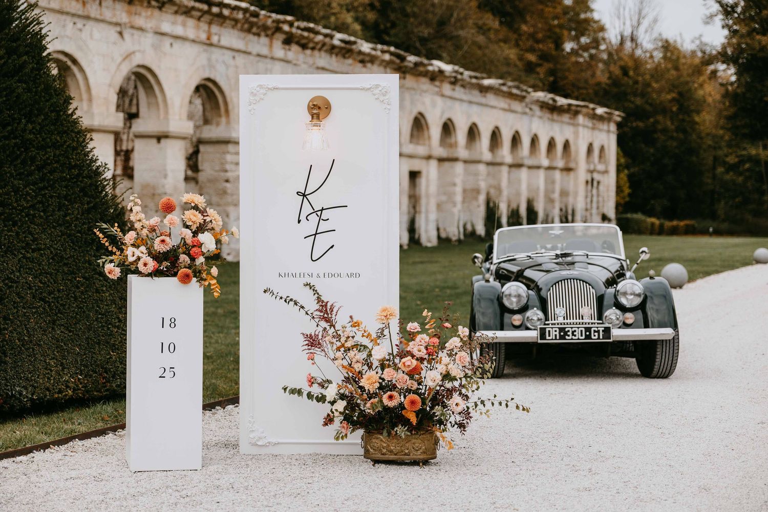 Elegant wedding welcome sign with vintage car and floral arrangements at chateau venue.