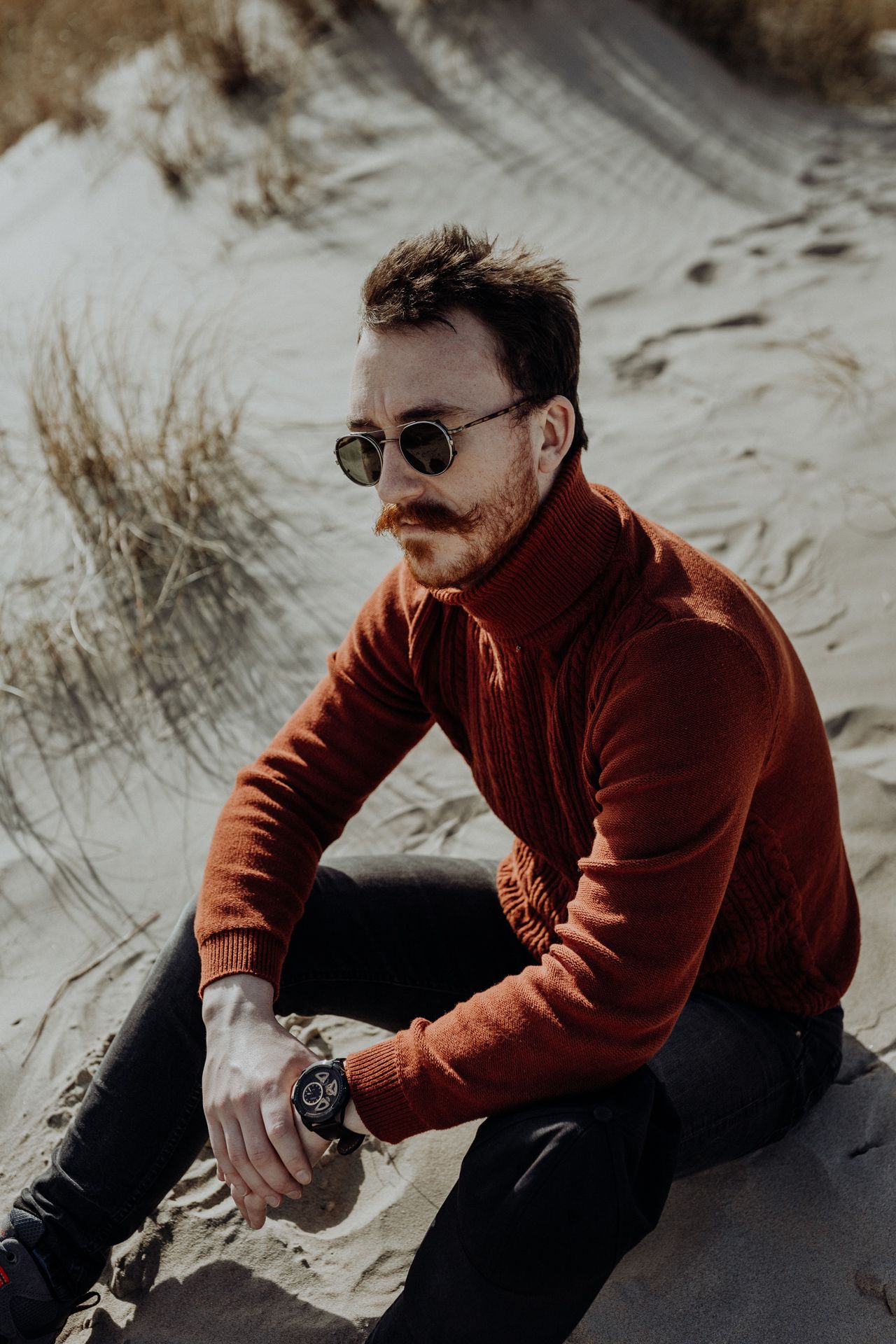 Stylish man with mustache wearing rust turtleneck and round sunglasses sitting on sand dunes.