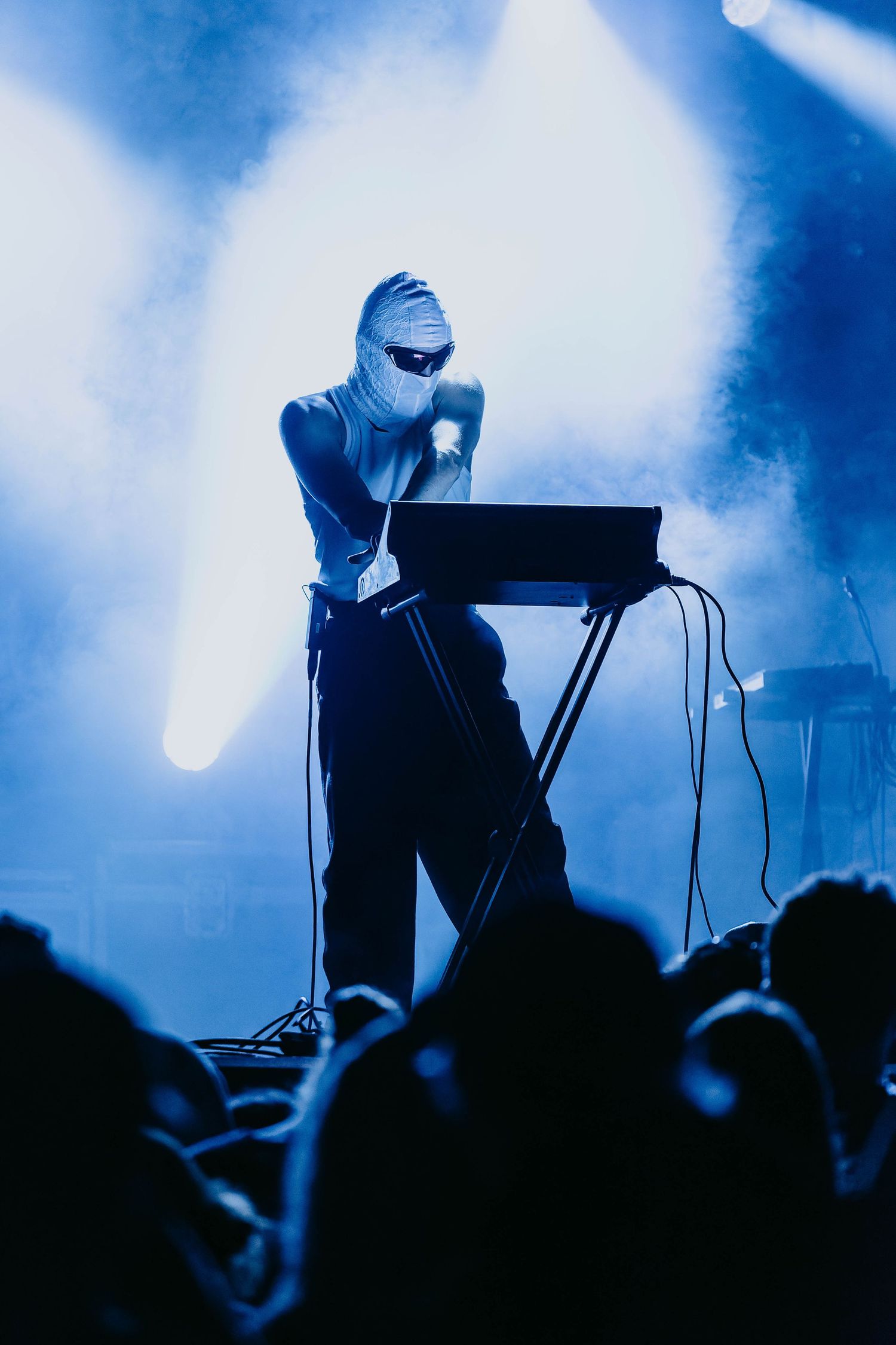 Masked performer singing at a keyboard on a blue smoke-filled concert stage before a crowd.
