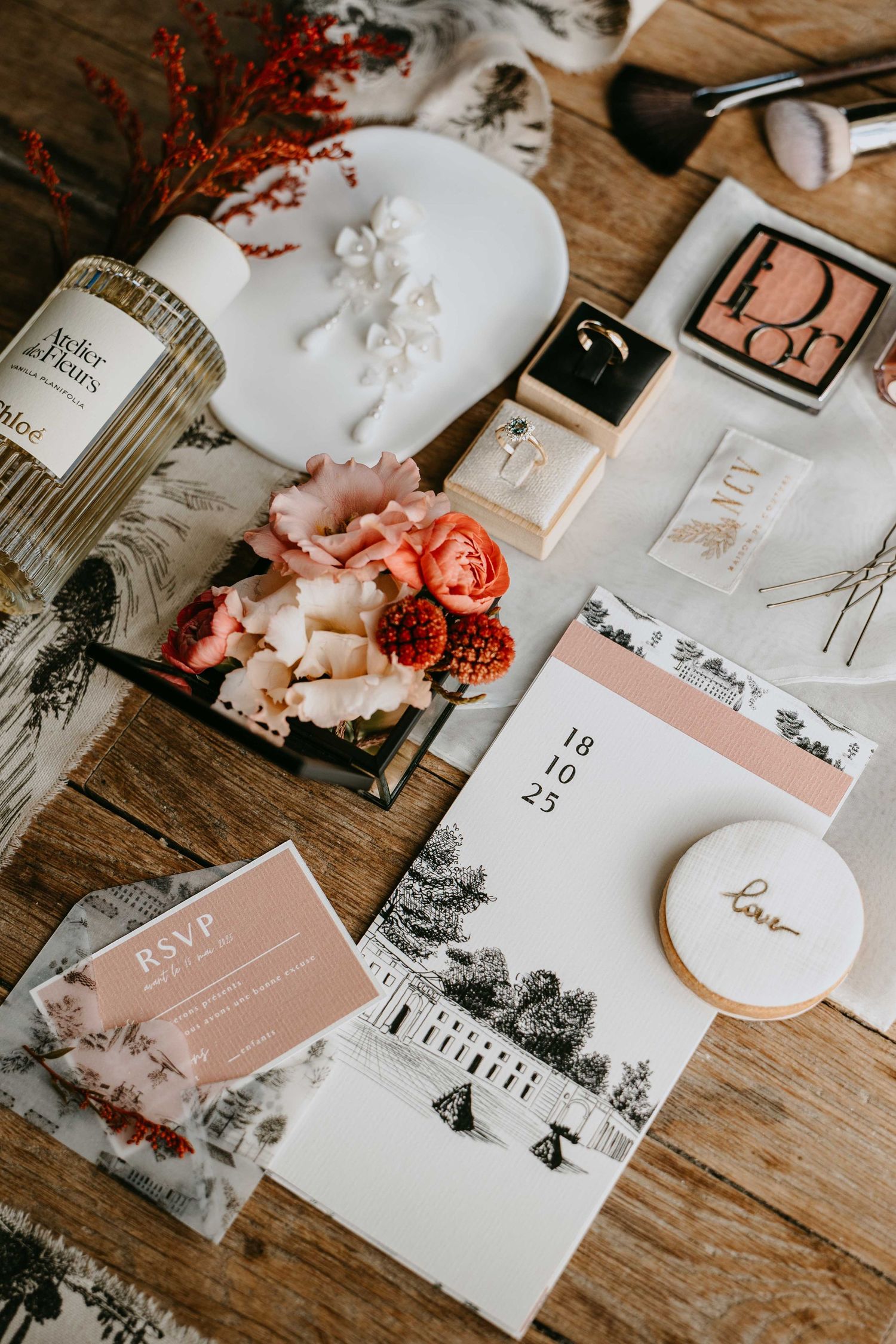 Elegant wedding flat lay with invitation suite, RSVP card, Dior makeup, floral arrangement, and jewelry on rustic wood.