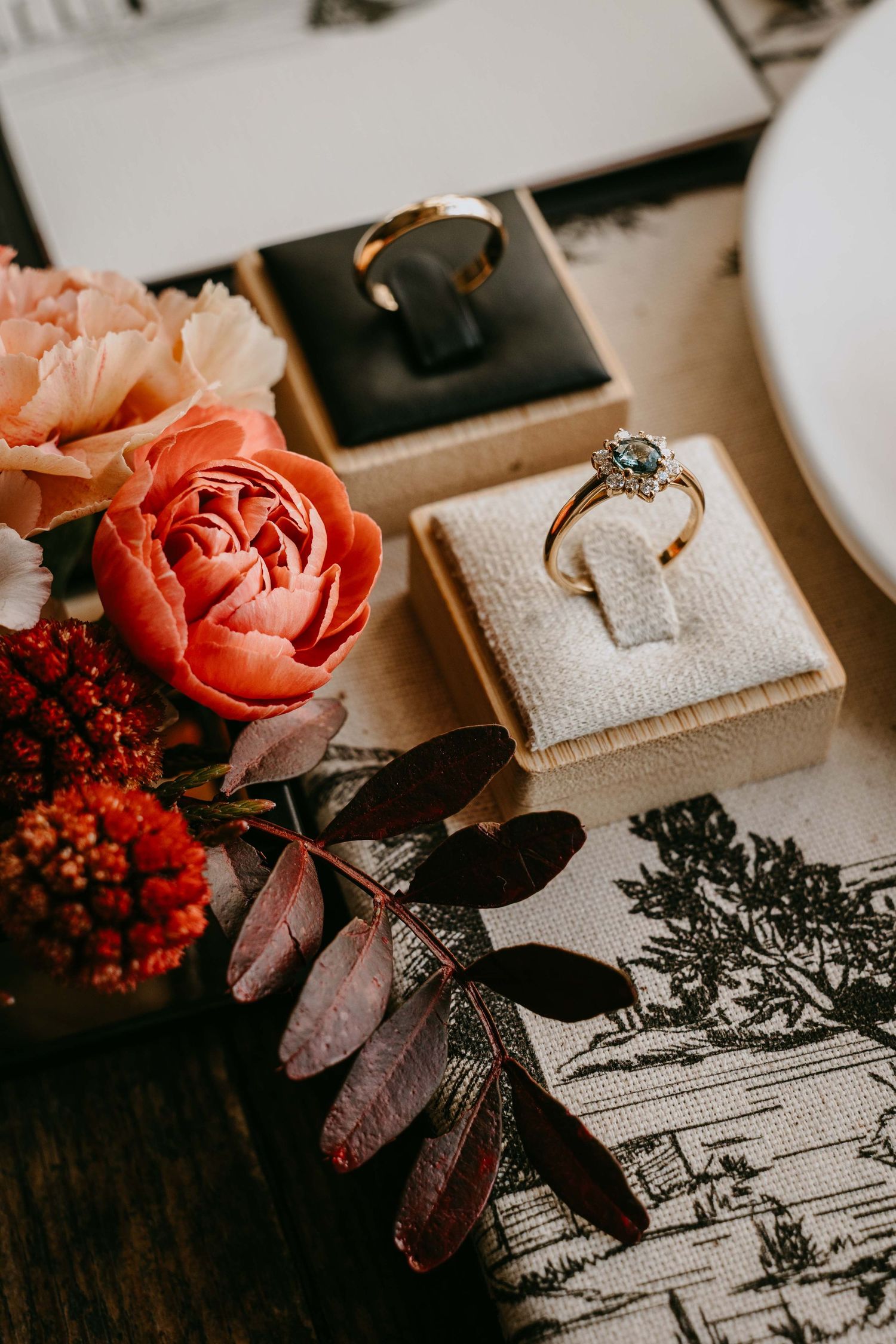 Gold engagement ring with diamond solitaire in cream velvet box beside coral roses and autumn leaves on toile fabric.