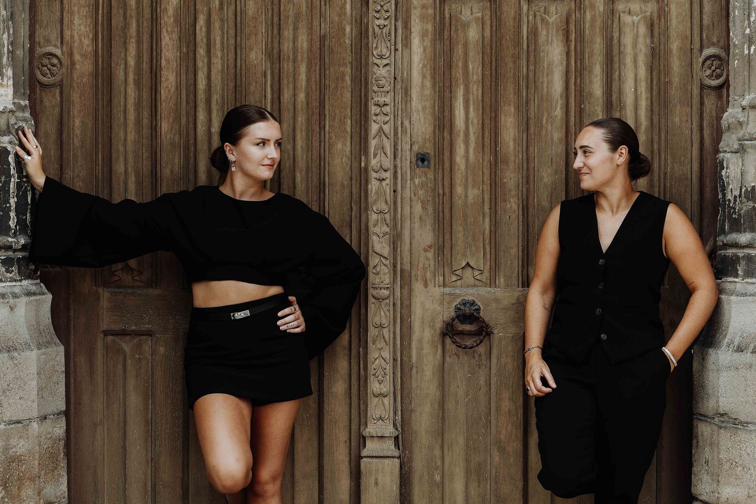 Two women dressed in all-black outfits posing against a rustic wooden door in an outdoor setting.