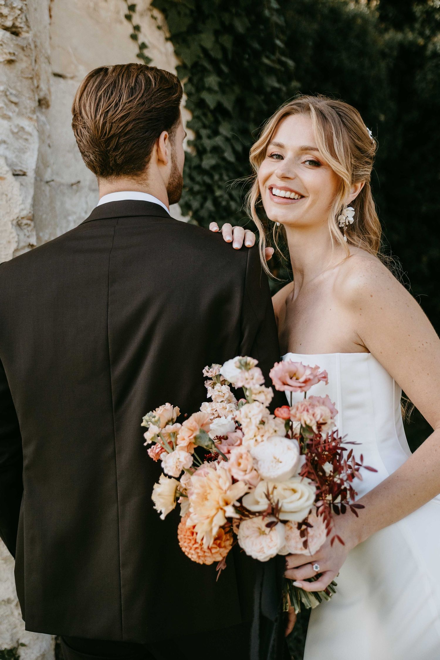 Smiling bride in strapless white gown holding a lush peach and pink floral bouquet beside her groom in a black suit.