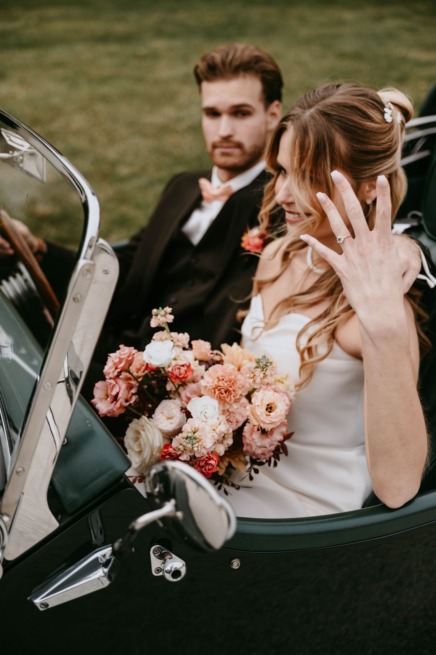 Bride shows off wedding ring while holding floral bouquet in vintage convertible car with groom seated behind her.