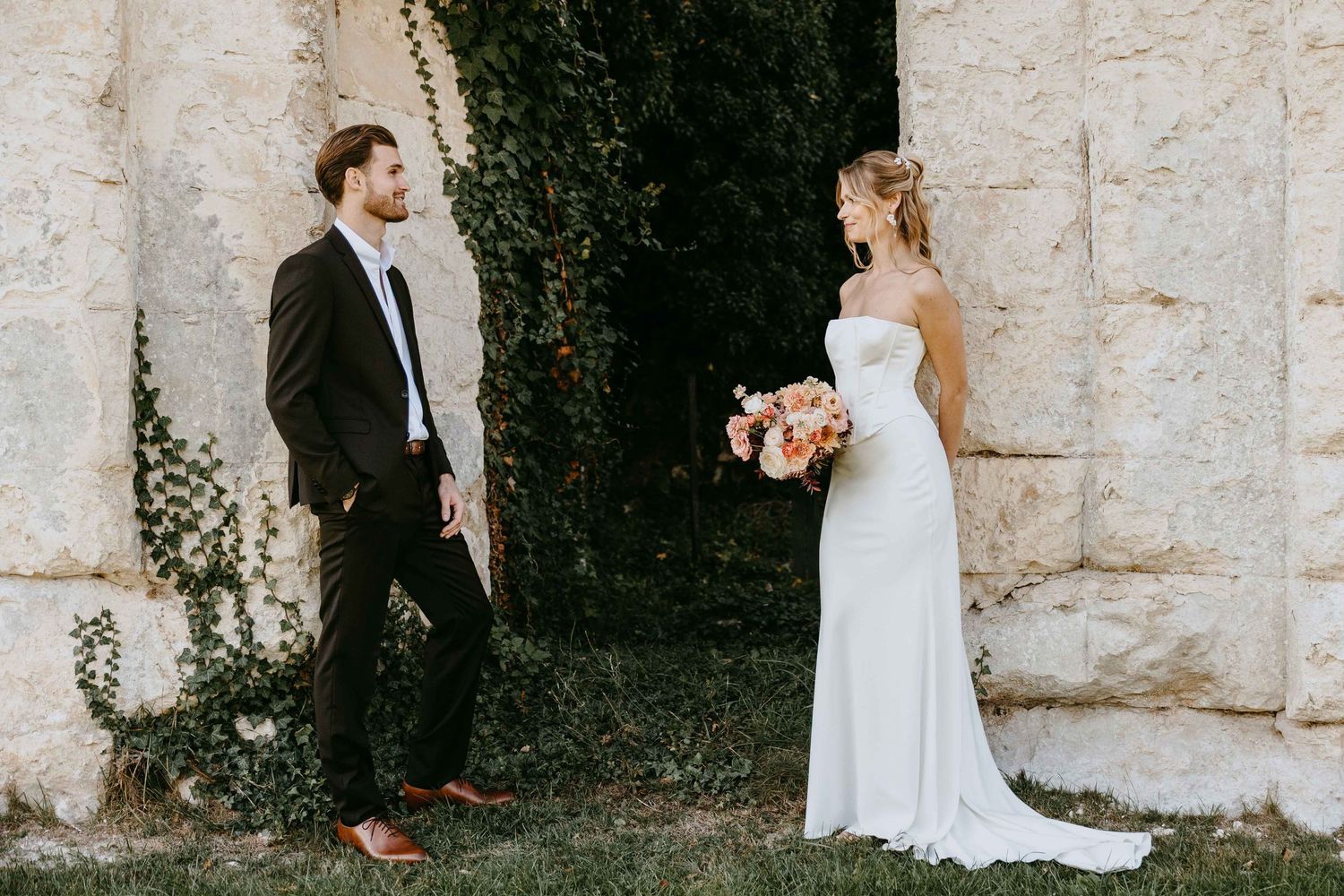 A bride in a white gown holding a bouquet faces a groom in a dark suit outdoors near a stone wall.