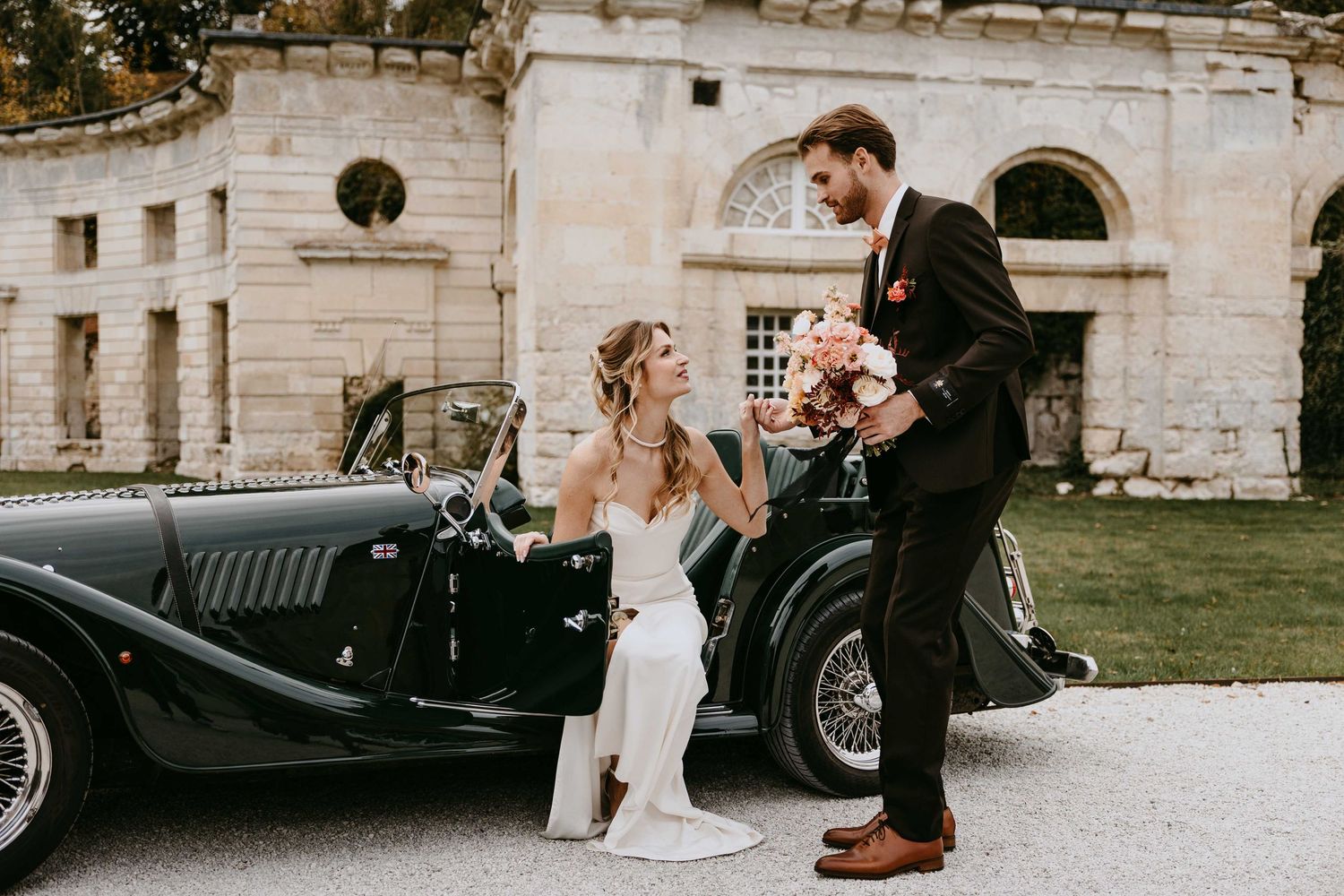 Bride and groom posing beside a vintage black convertible car outside a historic stone building on their wedding day.