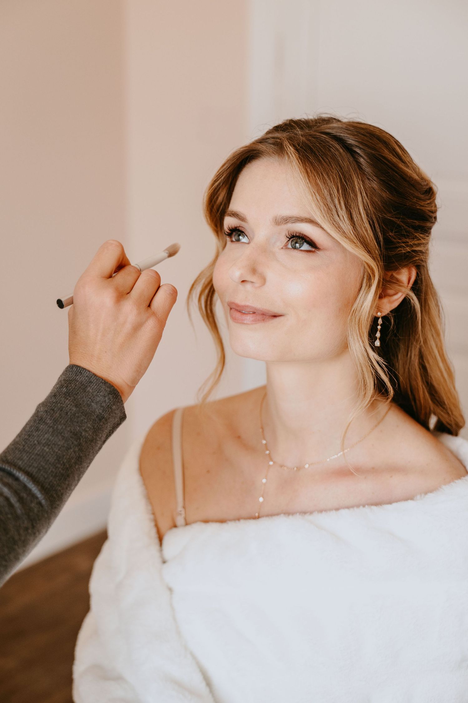 A makeup artist applying foundation to a smiling bride wearing a white dress and pearl jewelry.