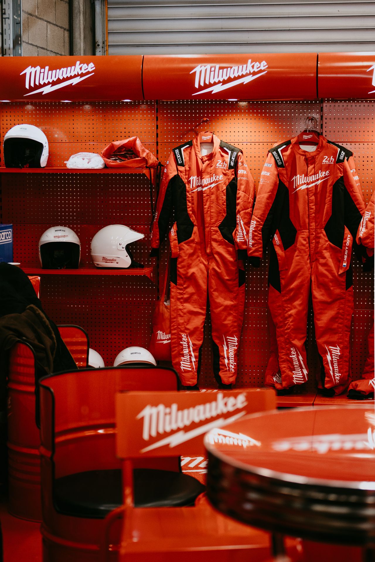 Milwaukee-branded red racing suits and helmets displayed on a pegboard wall in a race team garage.