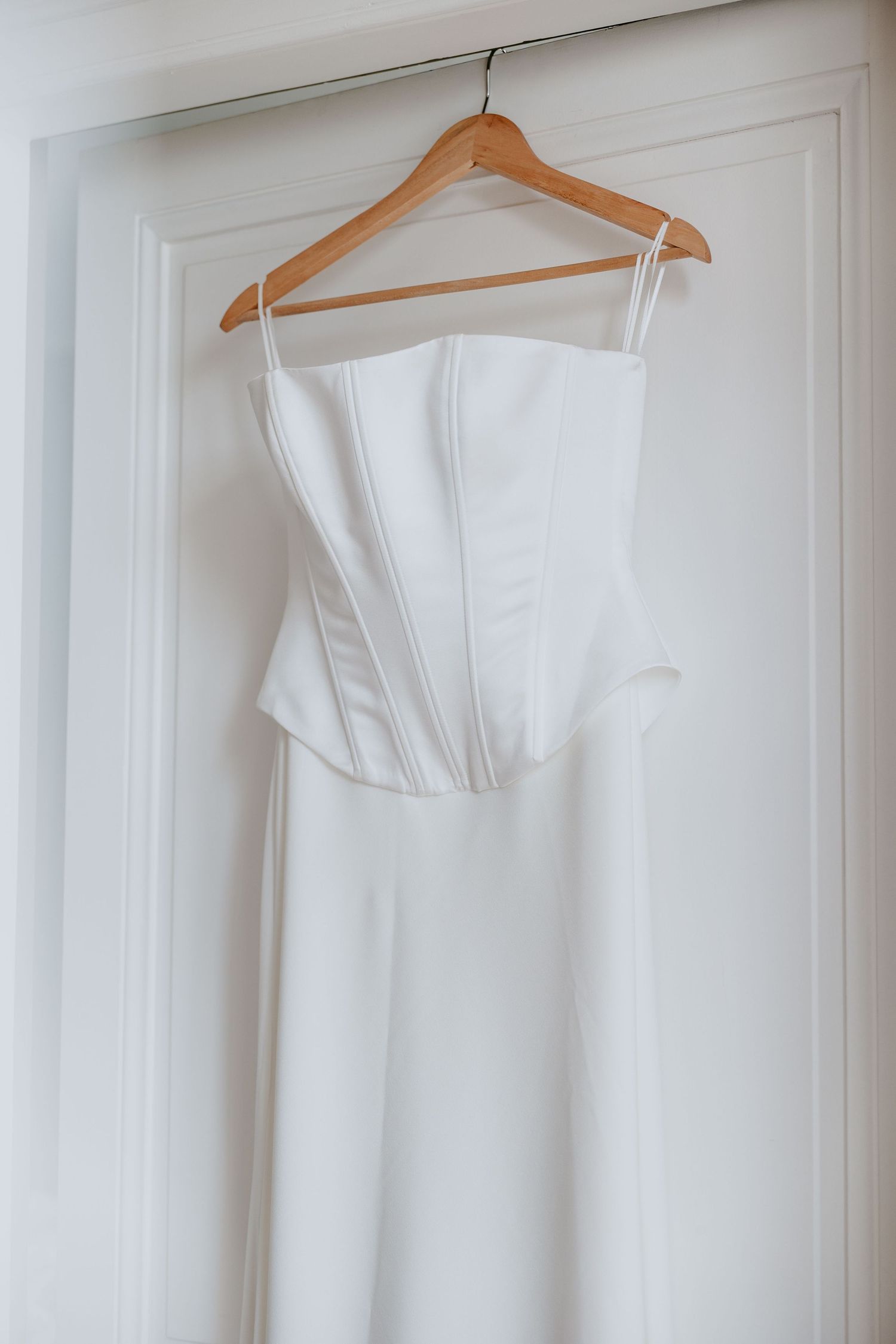 Minimalist white strapless corset wedding dress hanging on a wooden hanger against a white door.