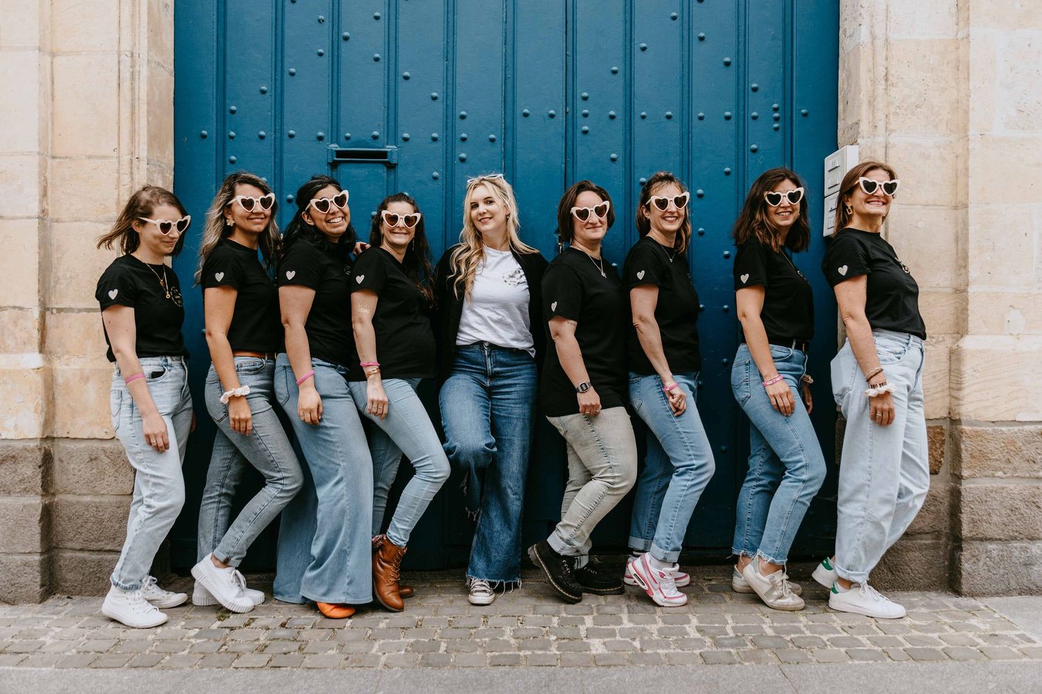 Row of women in matching black shirts and jeans leaning against a blue door outdoors.