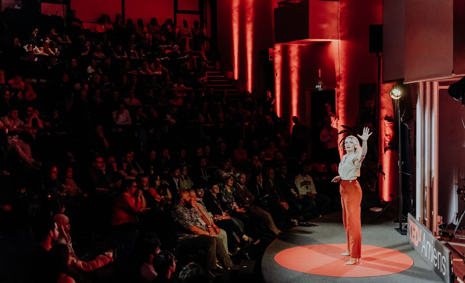 Speaker on stage in red-lit venue gestures to large seated audience during live event.