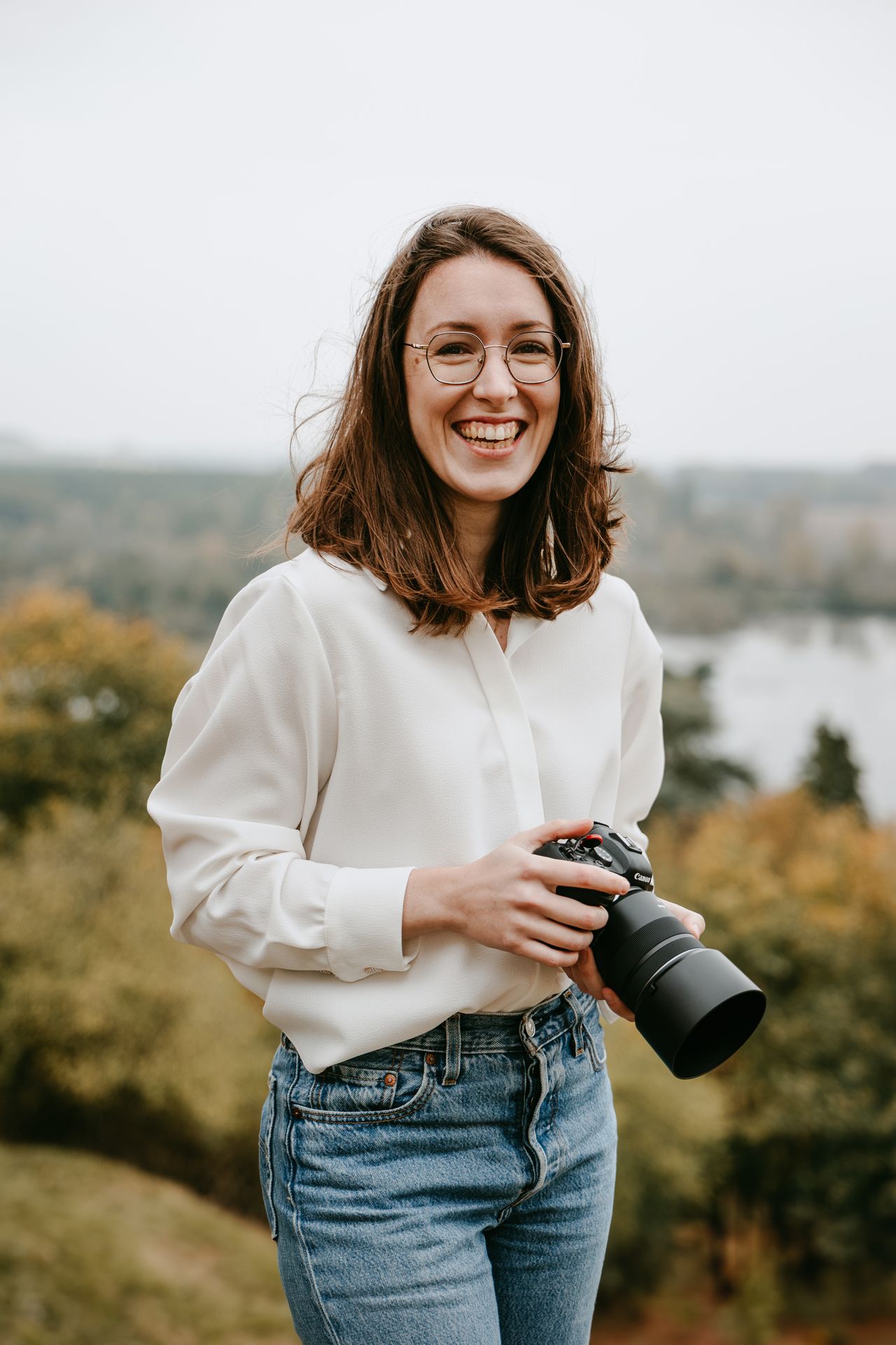 Smiling female photographer holding a DSLR camera outdoors with autumn foliage in the background.