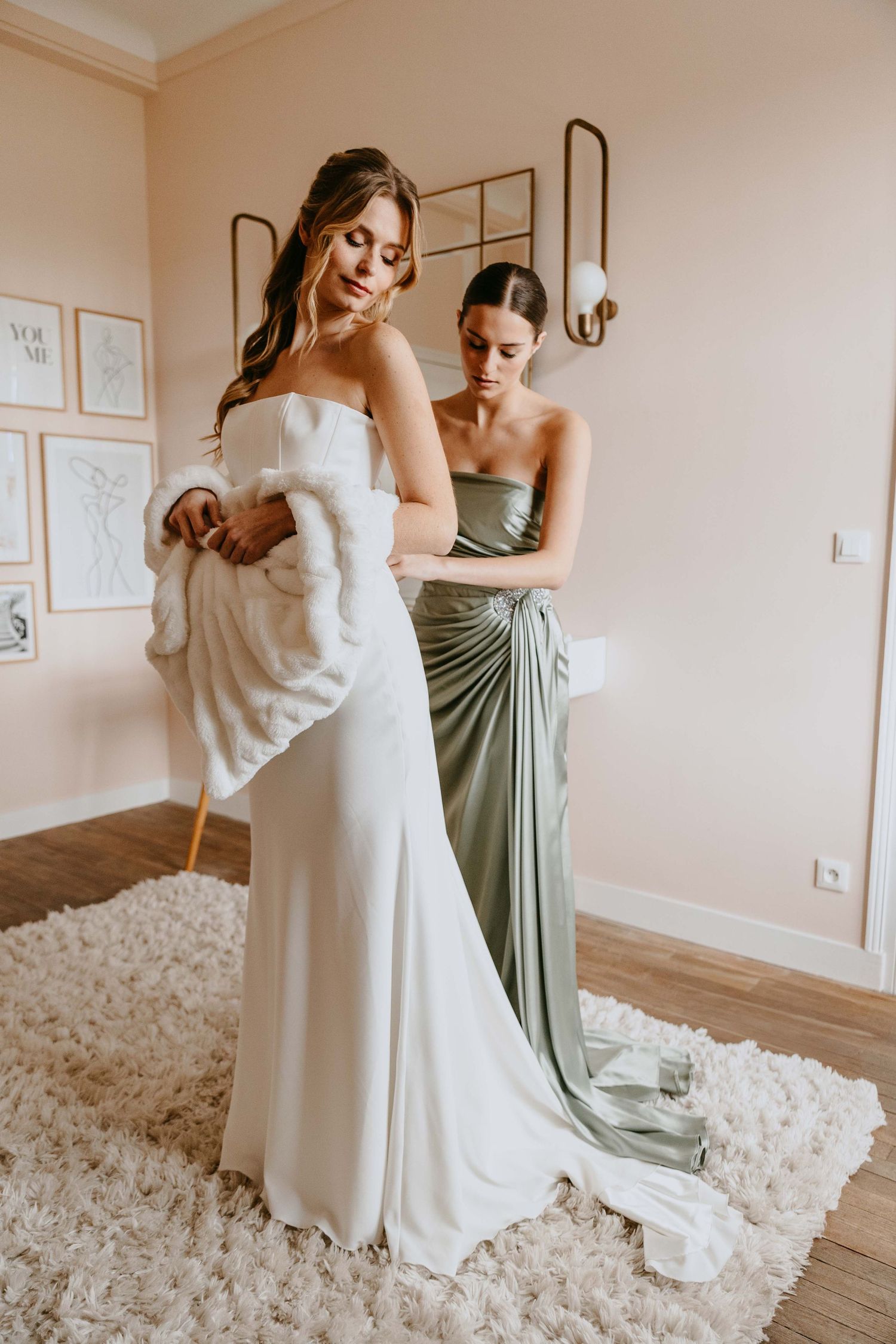 Bridesmaid in sage green dress helping bride in white gown put on fur wrap.