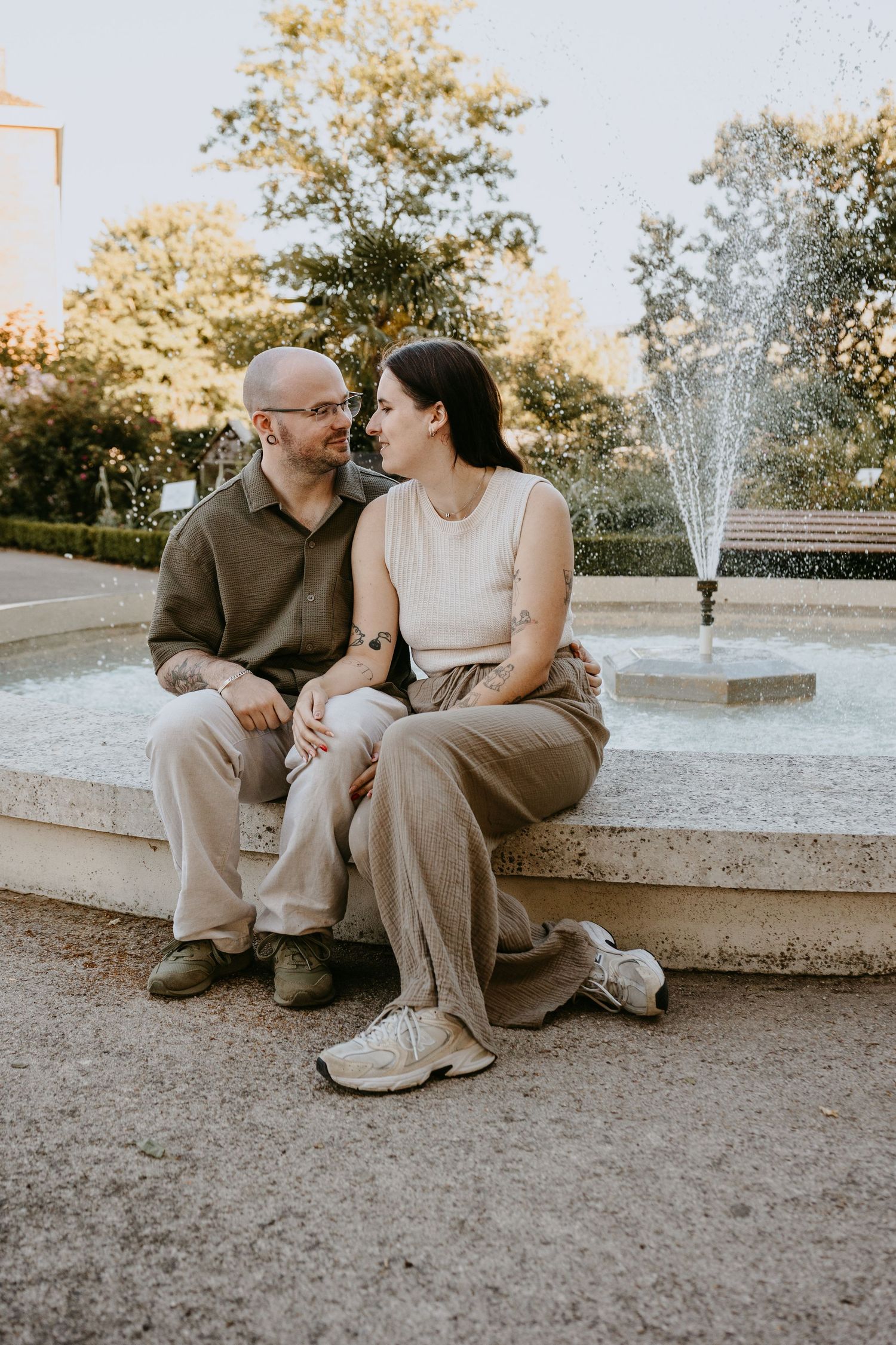 Couple sitting together by a fountain in a park, sharing a tender moment in neutral toned outfits.