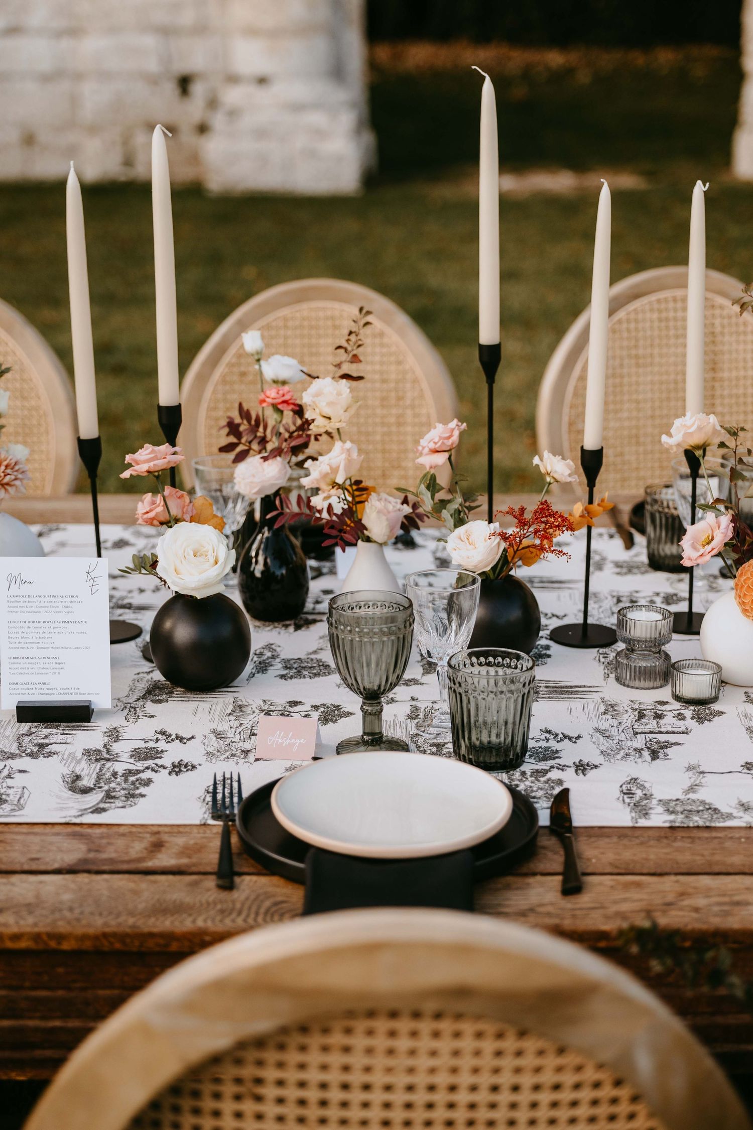 Elegant outdoor wedding tablescape with black candles, floral centerpieces, toile runner, and rattan chairs.