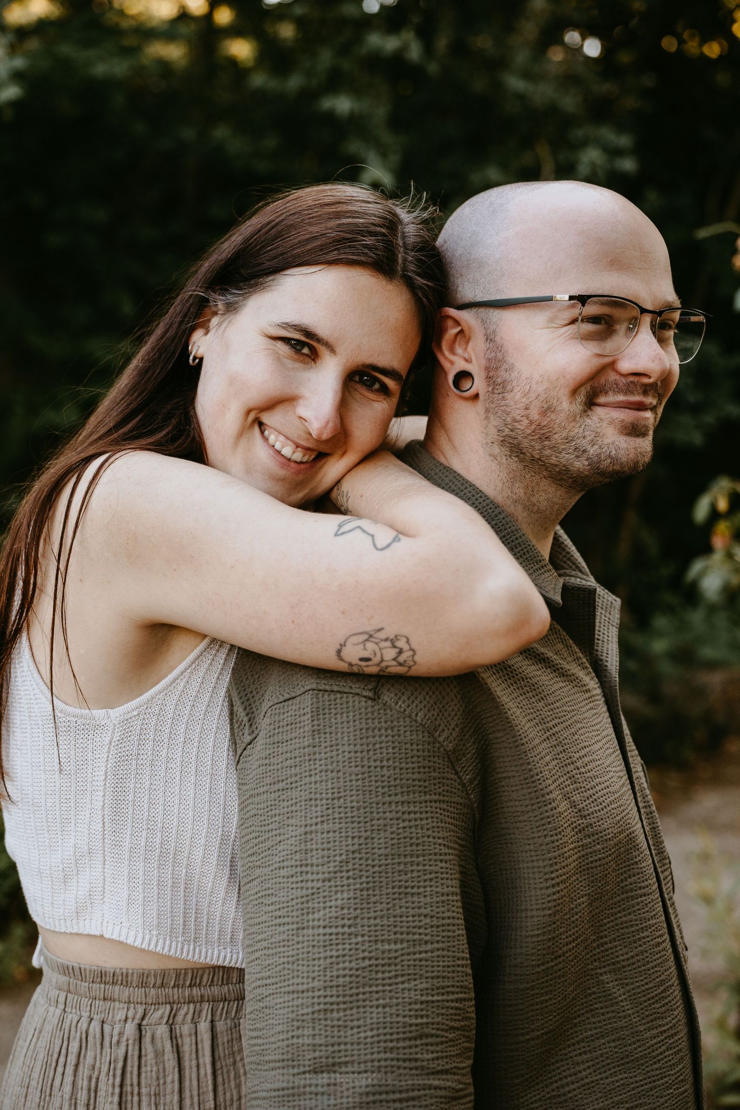 A smiling woman with tattoos hugs a bald man with glasses from behind outdoors near green trees.