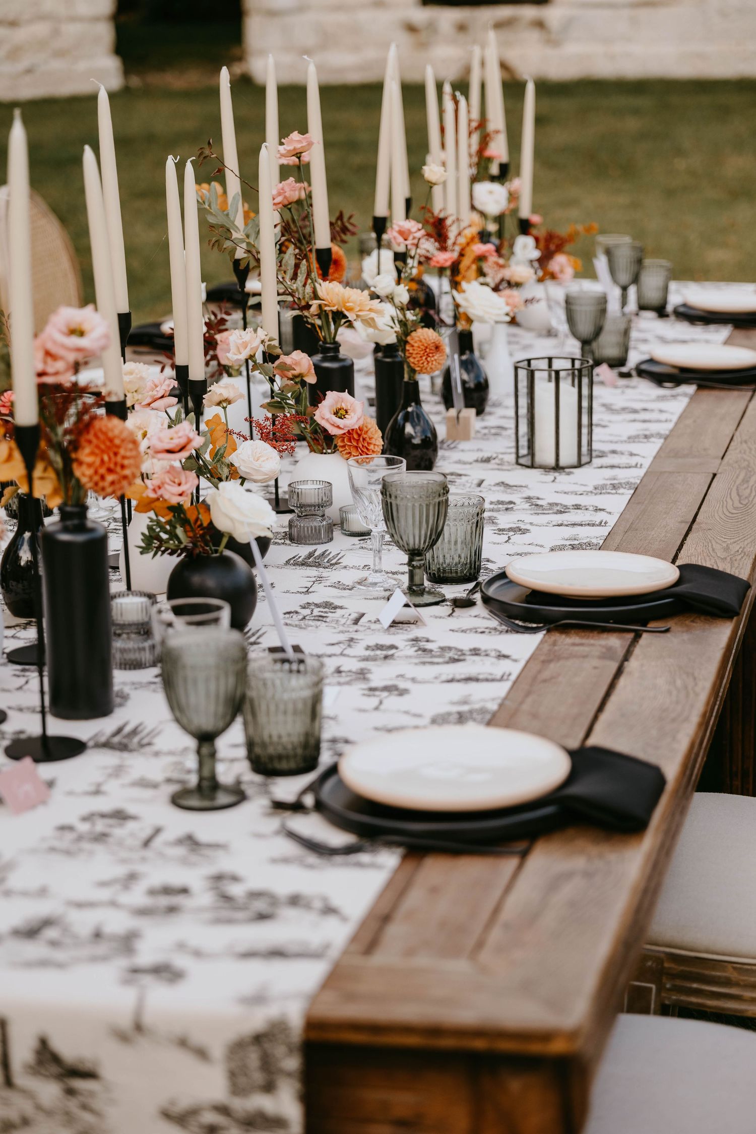 Elegant outdoor wedding tablescape with black candles, floral arrangements, gray goblets, and black plates on a rustic wood table.
