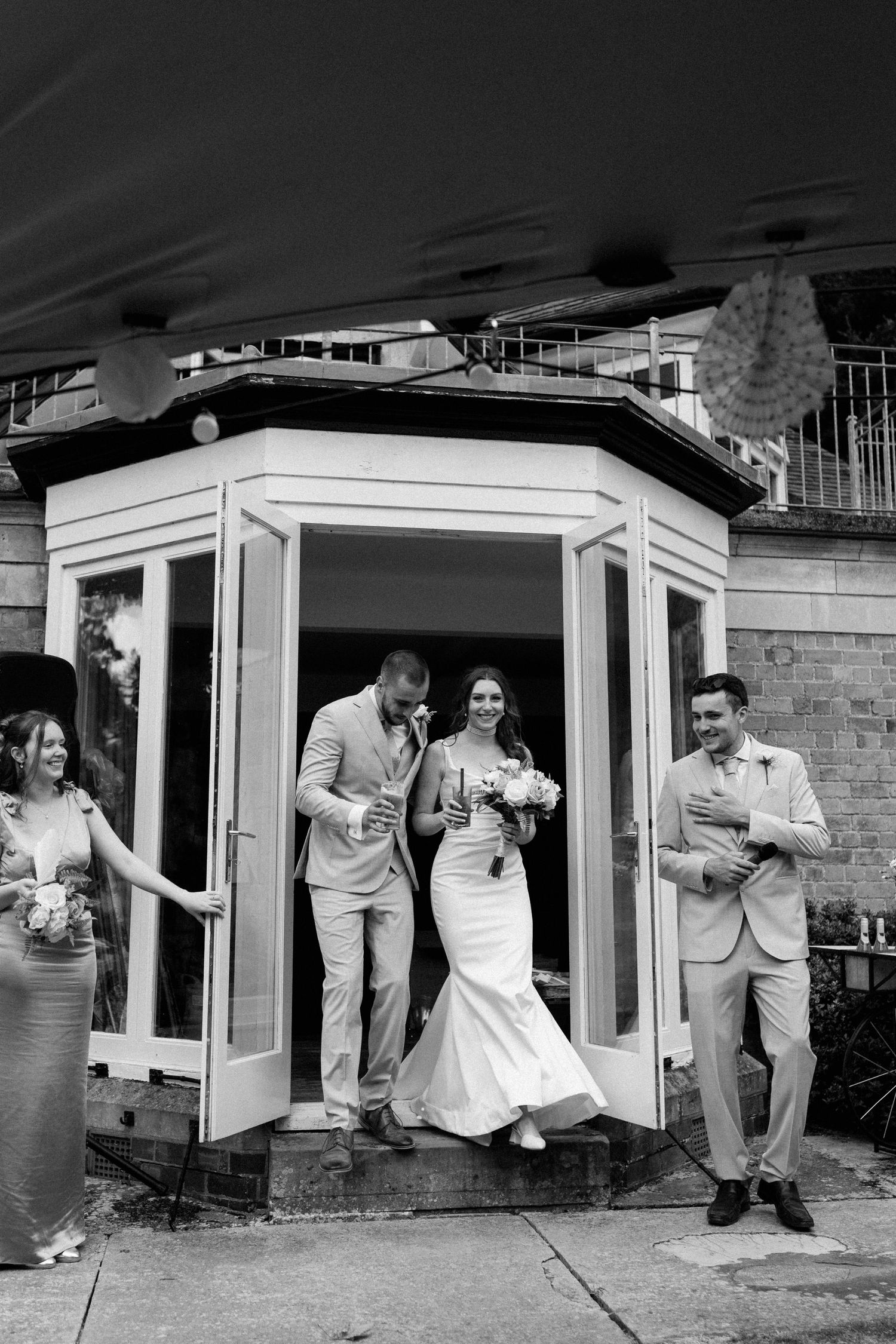 Bride and groom exit venue doors in black and white photo, flanked by wedding party holding champagne glasses.