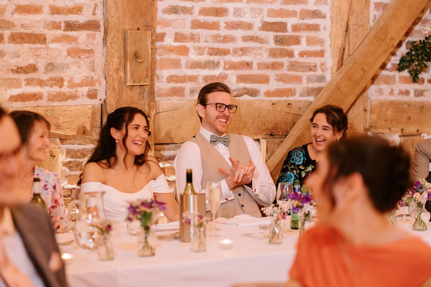 Bride and groom laughing at wedding reception table in rustic brick barn with floral centerpieces.