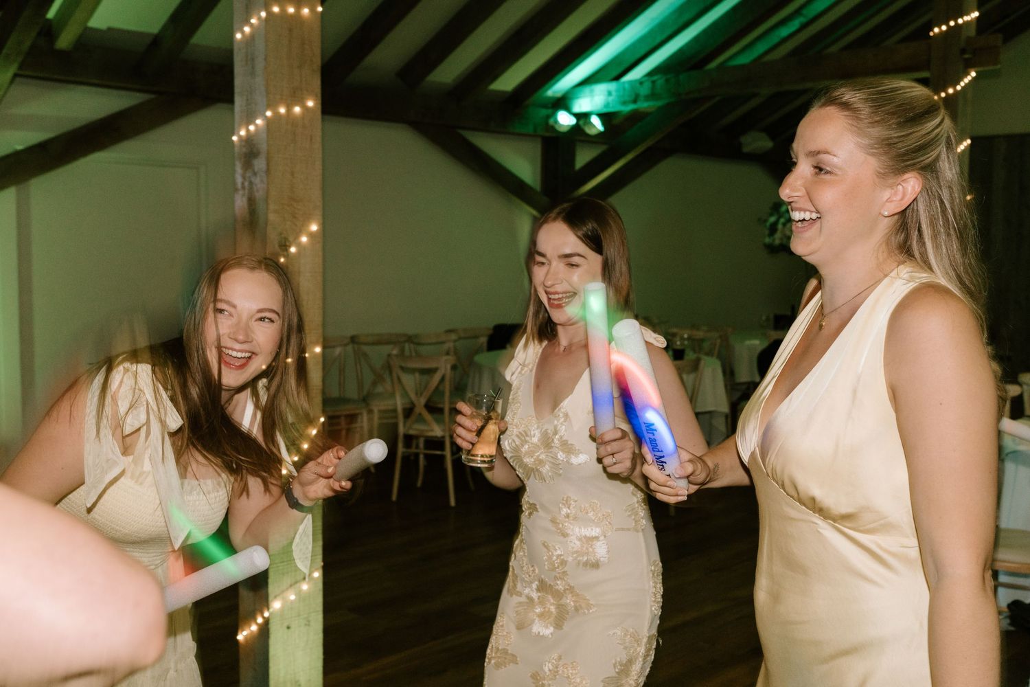 Three women in formal dresses laugh and dance with colorful glow sticks at a wedding reception.
