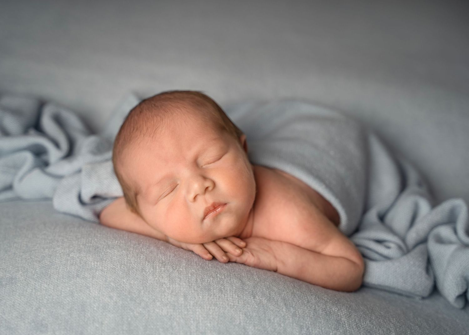 Peaceful newborn baby sleeping on a soft blue blanket, with tiny hands tucked under chin in a serene portrait.