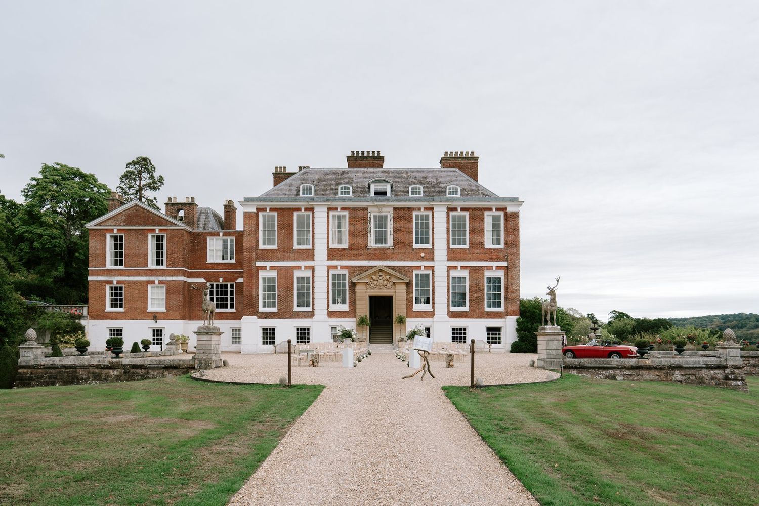 Grand red brick English country manor house with gravel driveway, manicured lawns, and elegant Georgian architecture under cloudy skies.