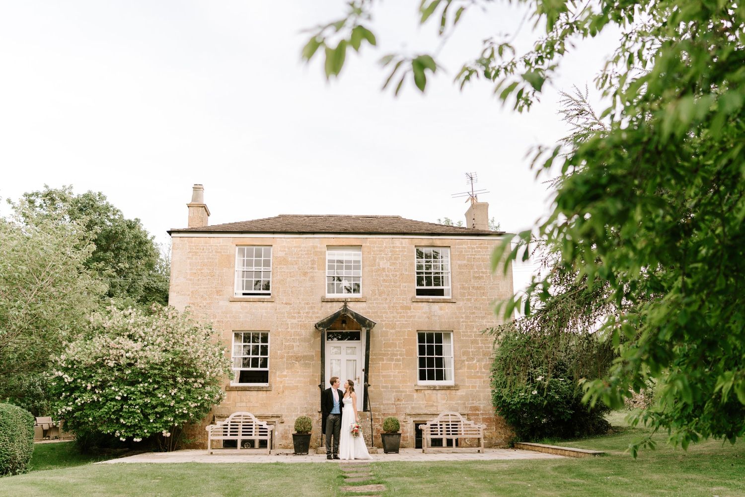 Bride and groom standing outside a beautiful two-story brick country house surrounded by lush greenery on their wedding day.