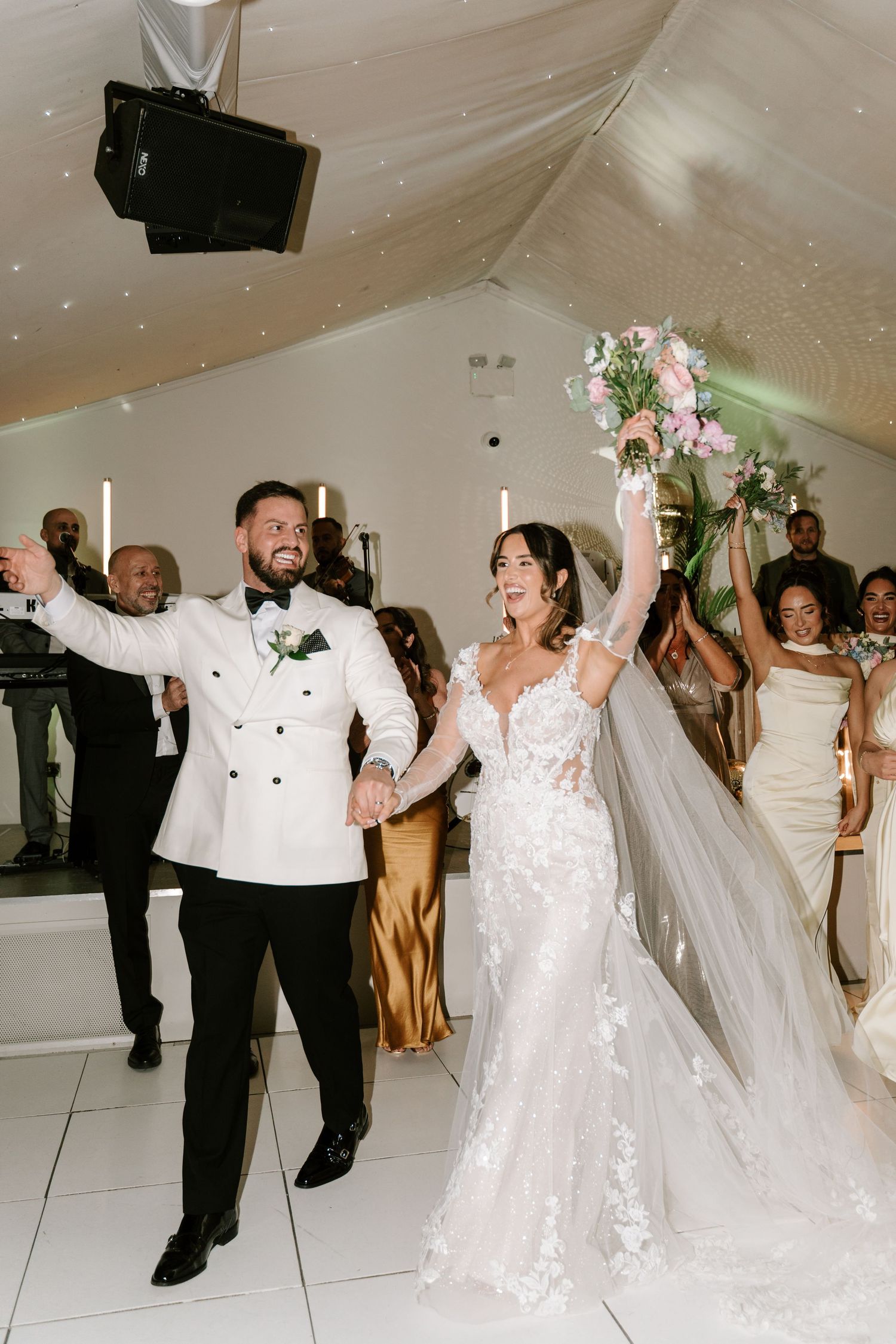 Bride and groom celebrate their first dance, she raises bouquet in lace gown with veil, he wears white tuxedo jacket.