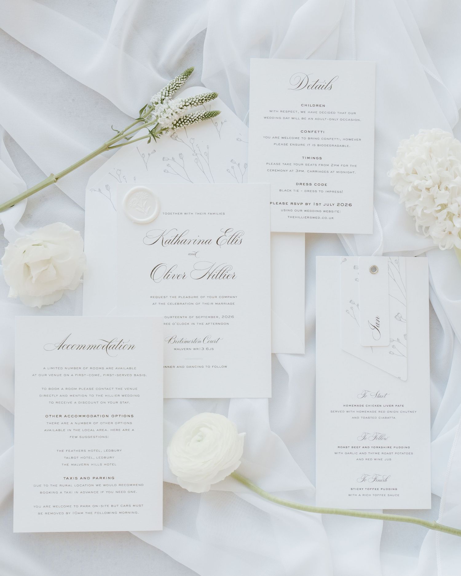 Elegant white wedding invitation suite with calligraphy script, wax seal, and floral accents on soft draped fabric.