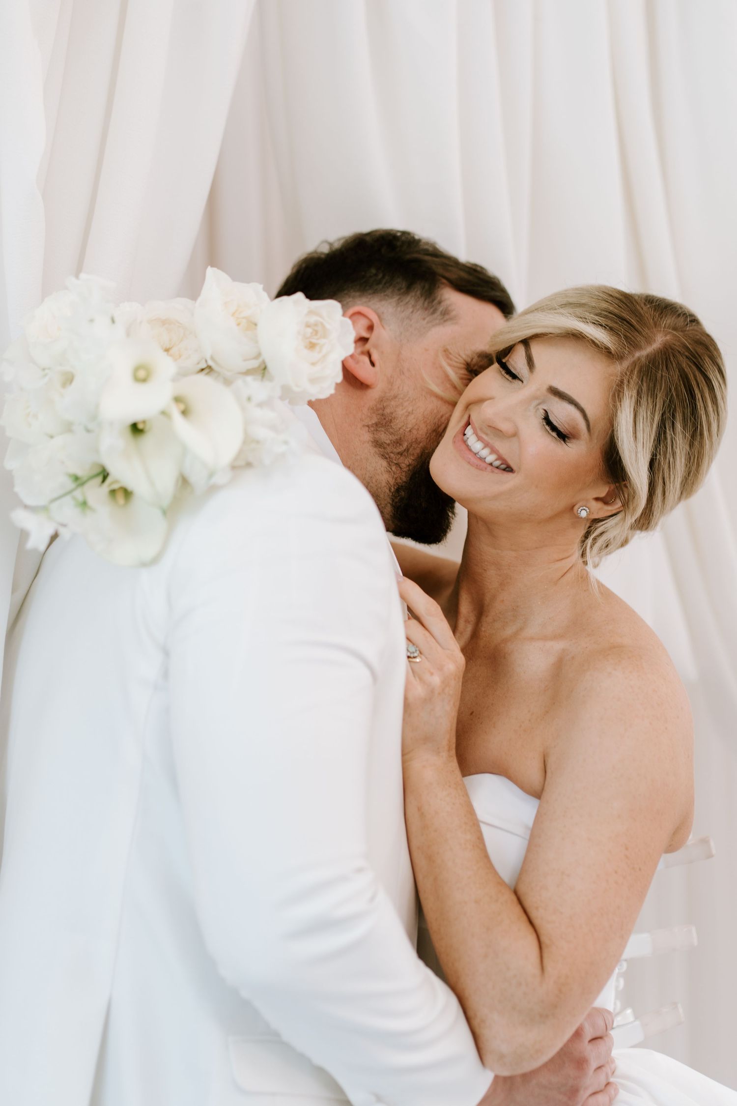 Bride smiling joyfully as groom kisses her cheek, both dressed in white, holding a white floral bouquet.