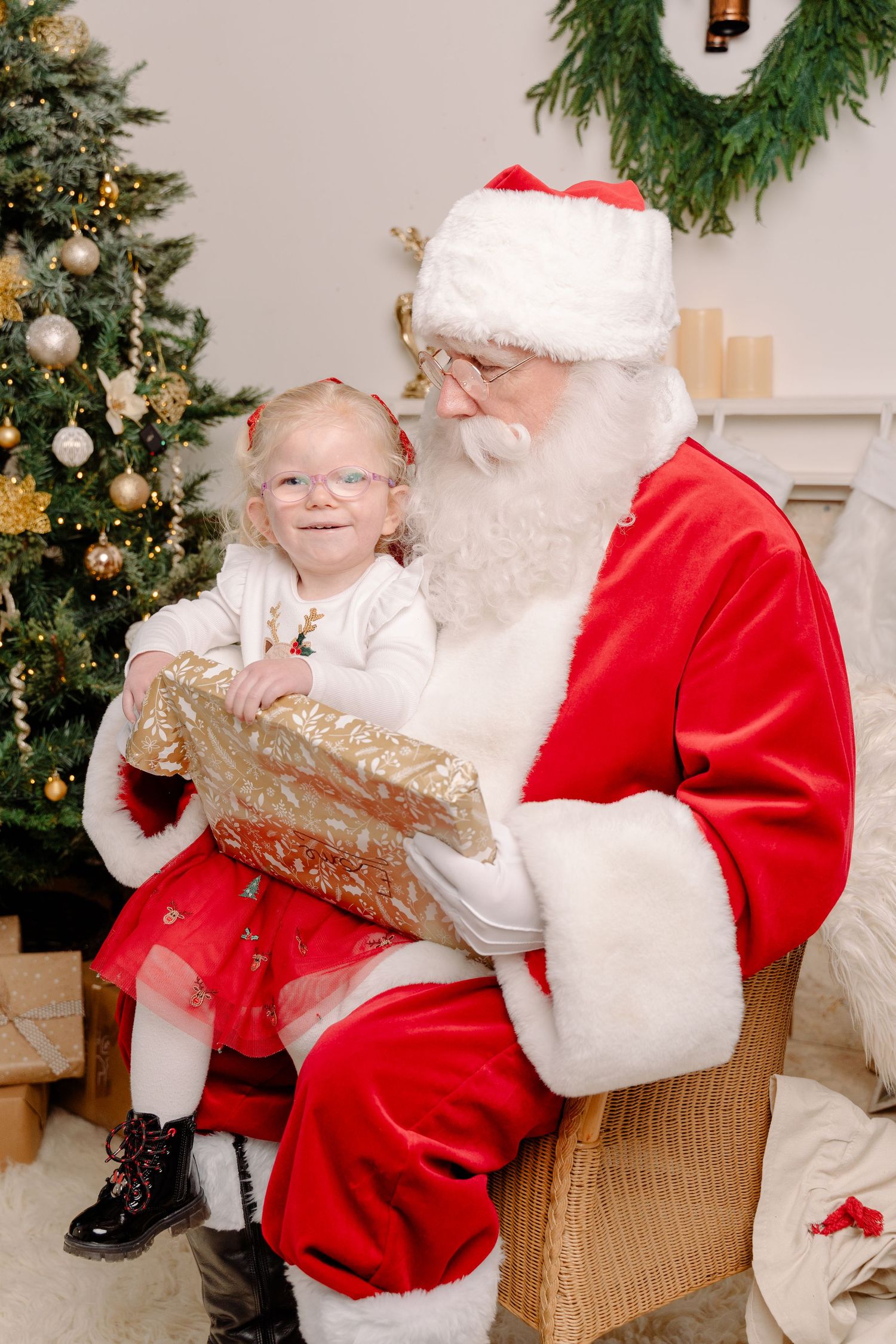 Young girl with glasses sits on Santa Claus lap holding a gold gift box near a decorated Christmas tree.