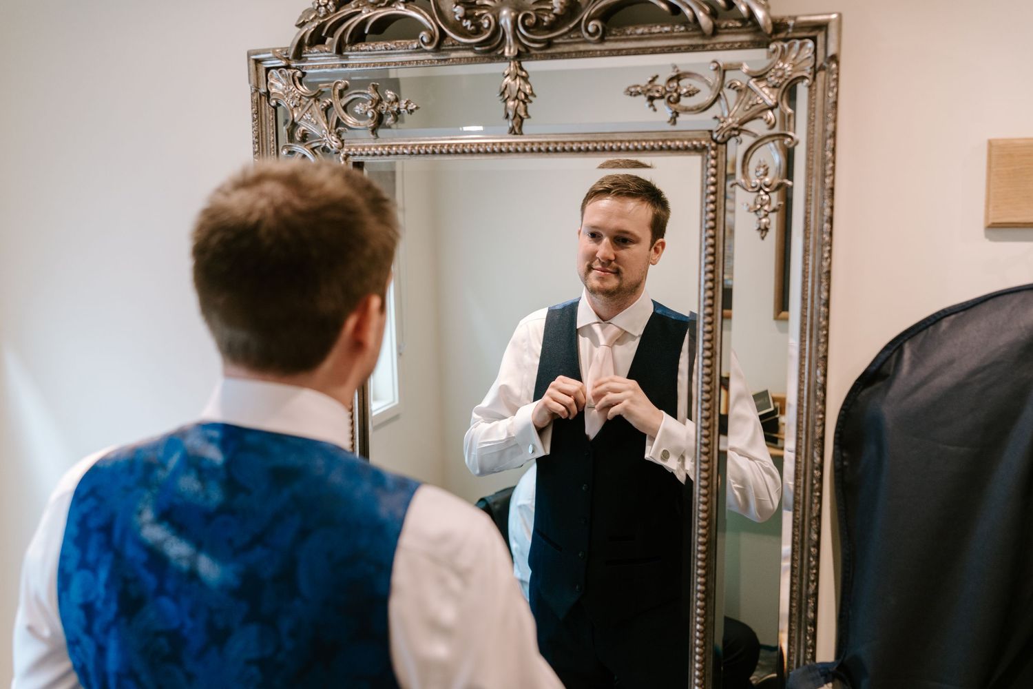 Groom adjusting his vest while getting ready, reflected in an ornate silver mirror before his wedding.