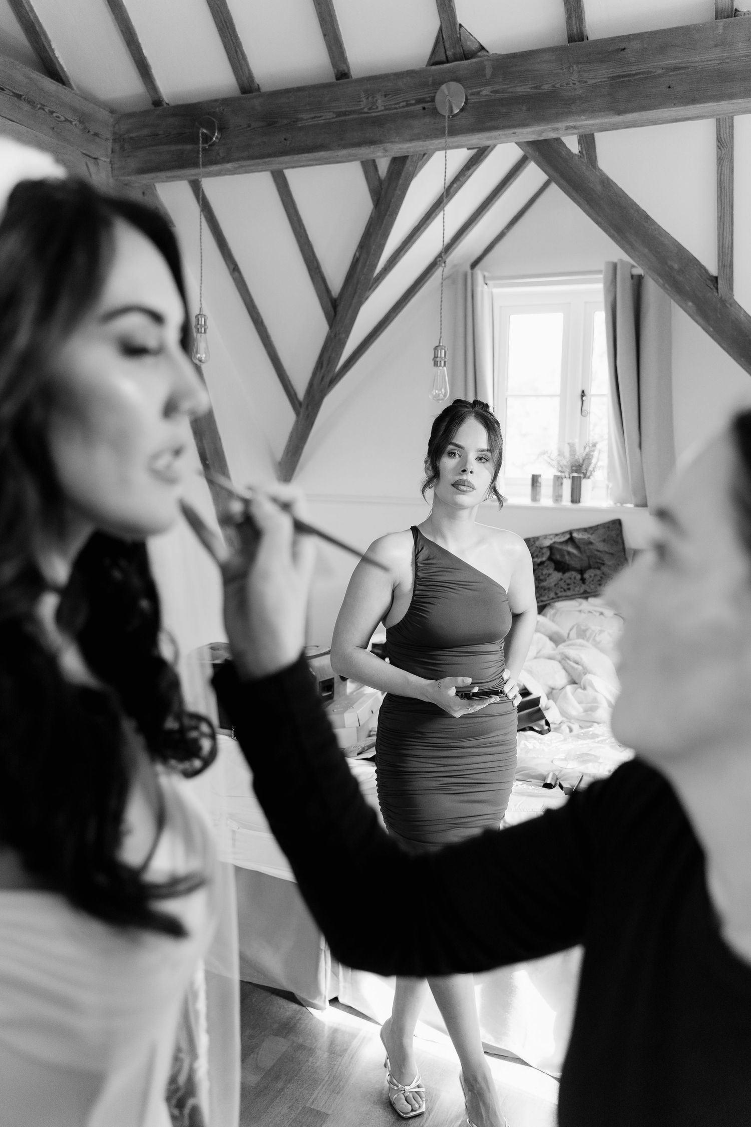 Black and white photo of a bride having makeup applied while a bridesmaid in a fitted dress watches in a rustic venue.